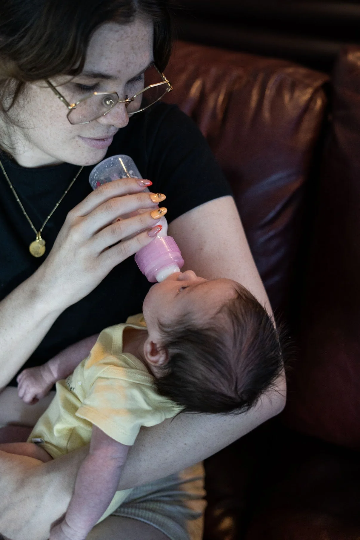 A woman with glasses and freckles is feeding a baby with a bottle while sitting on a brown leather couch.