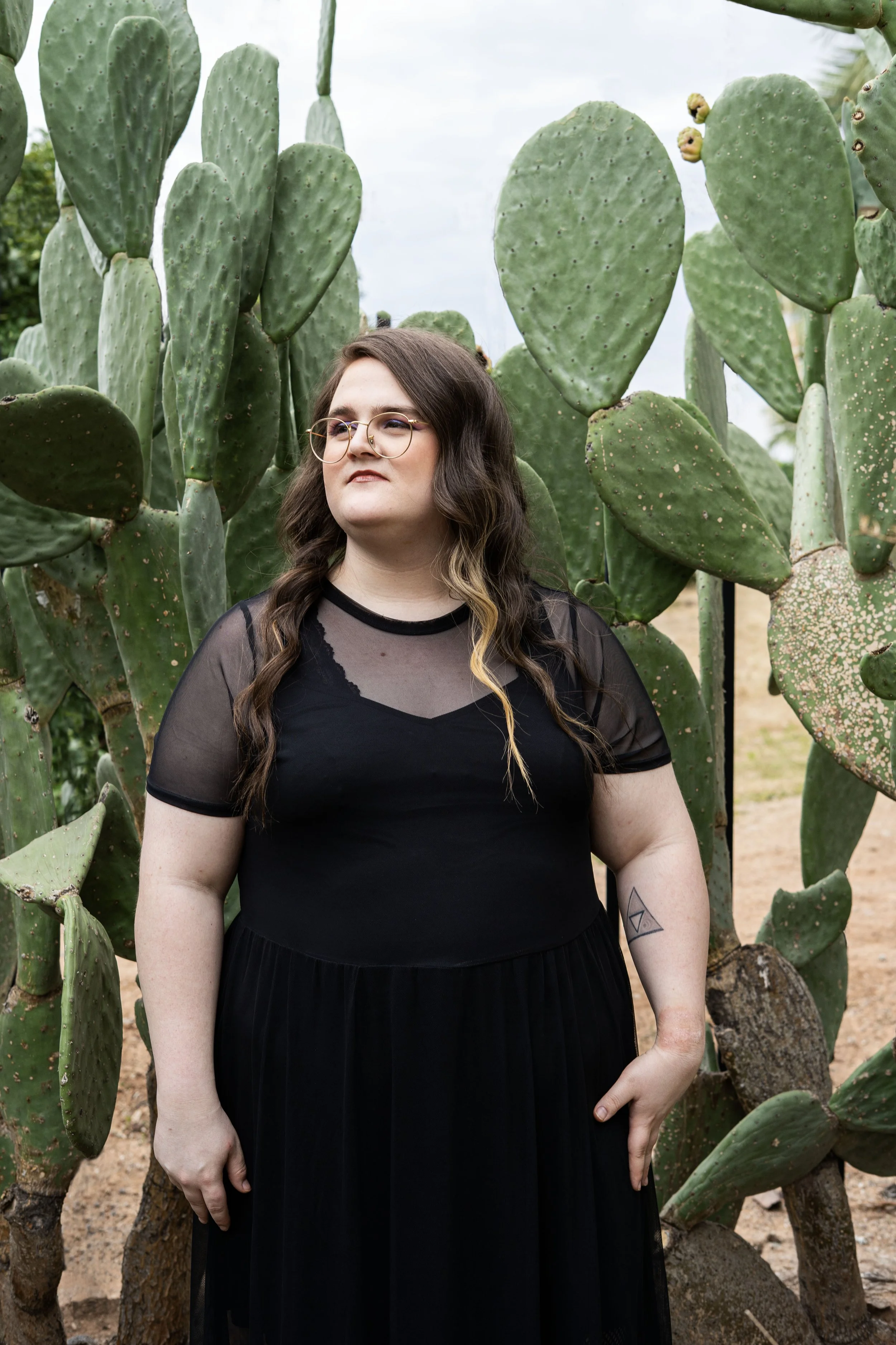 A woman with long brown hair, glasses, and a black dress standing among large prickly pear cactus plants outdoors during daytime.