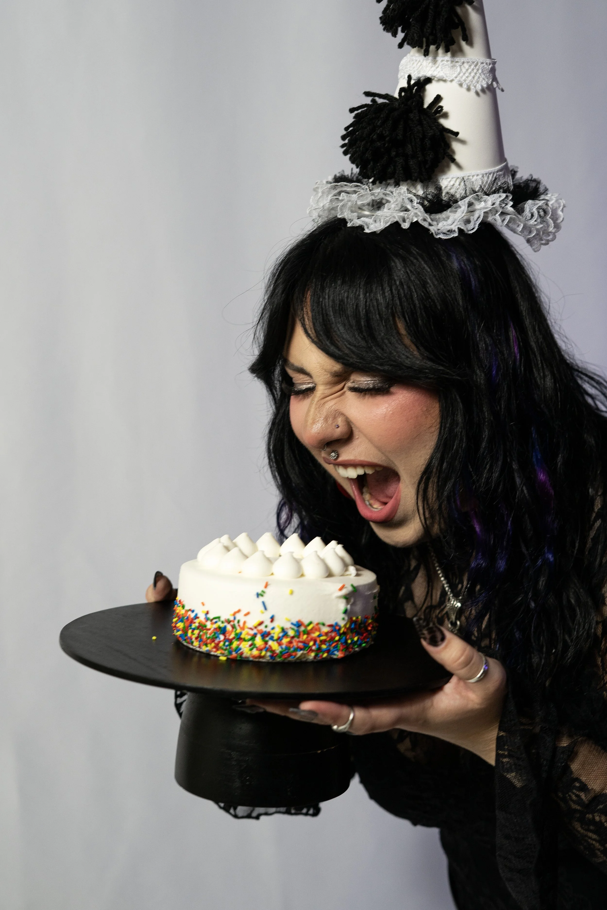 A woman with black hair, wearing a ruffled birthday hat with pom-poms, smiling and pretending to bite into a small birthday cake with rainbow sprinkles, holding it on a black platter.