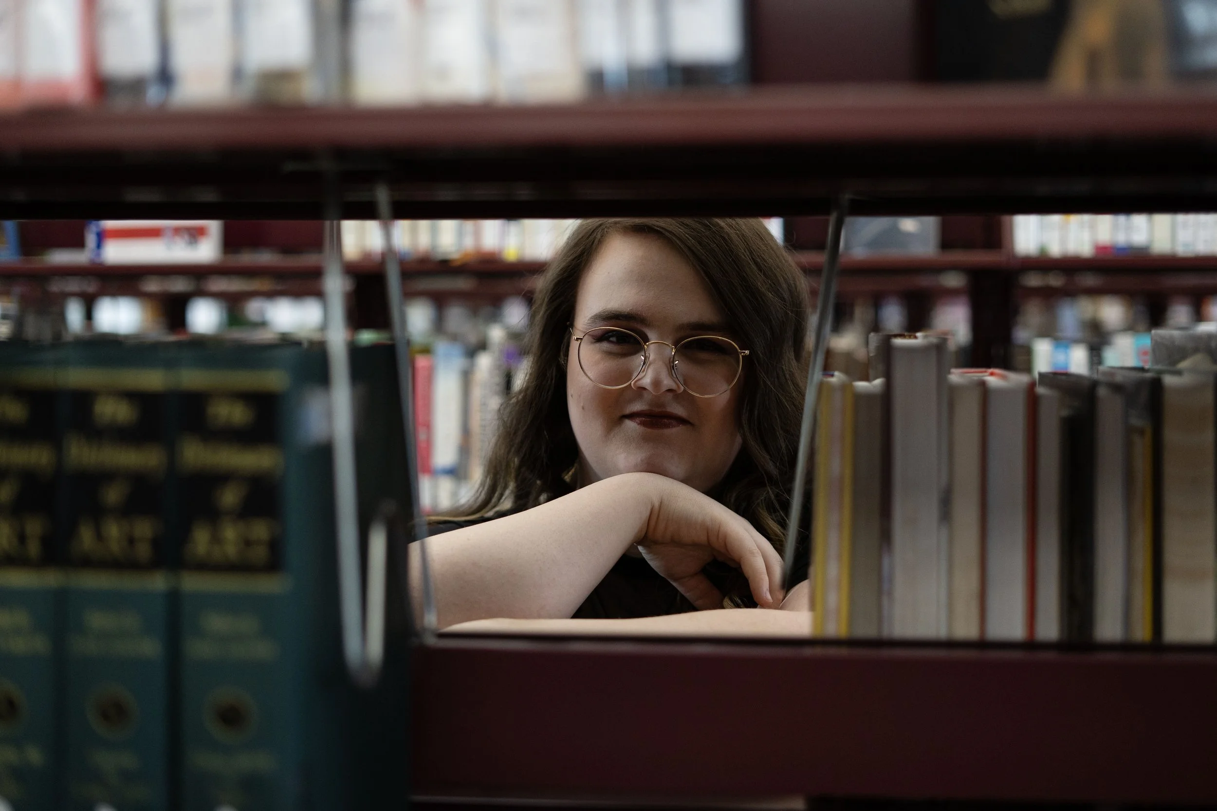 A woman with glasses and long brown hair is sitting at a bookshelf in a library, resting her chin on her arm and looking at the camera.