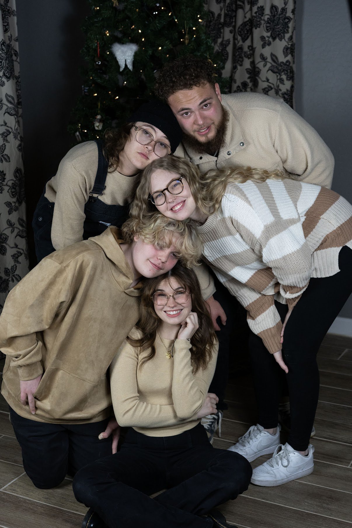 A group of six young people, posing in front of a decorated Christmas tree, smiling and dressed casually.
