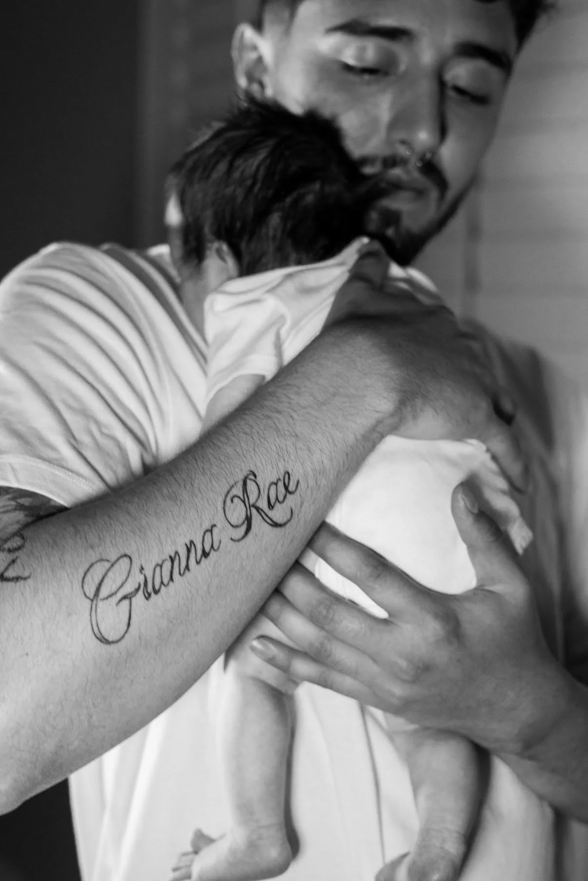 A man is holding a small child, cradling the child in his chest, with his eyes closed. The man's arm has a tattoo that says "Gianna Rae." The image is in black and white.