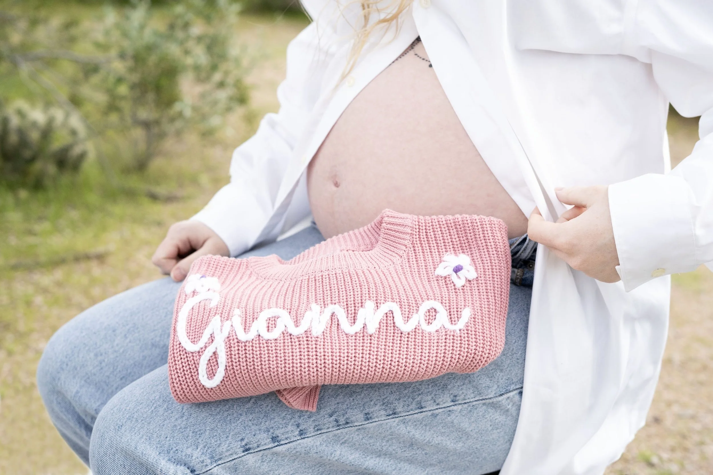 Pregnant woman holding pink knit blanket with white embroidery reading 'Gianna' and sitting outdoors.