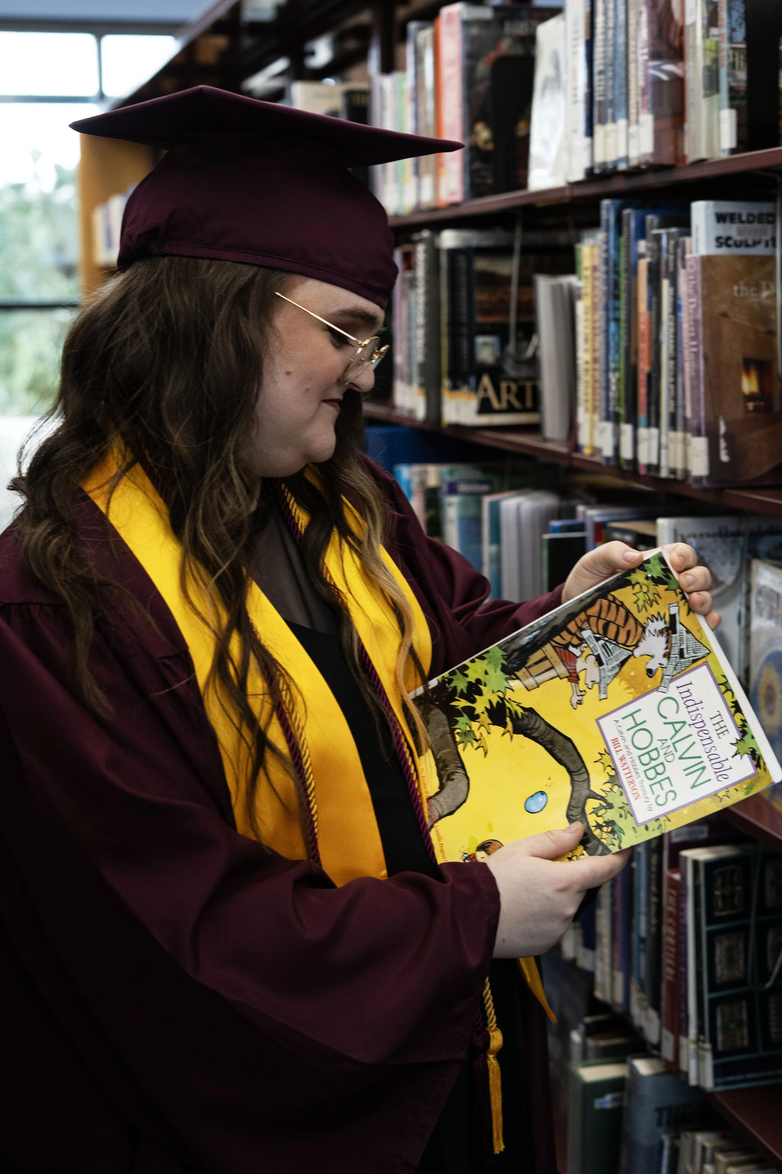 A young woman in a maroon graduation cap and gown, with a yellow stole, is looking at a book titled 'The Indispensable Calvin and Hobbes' in a library.