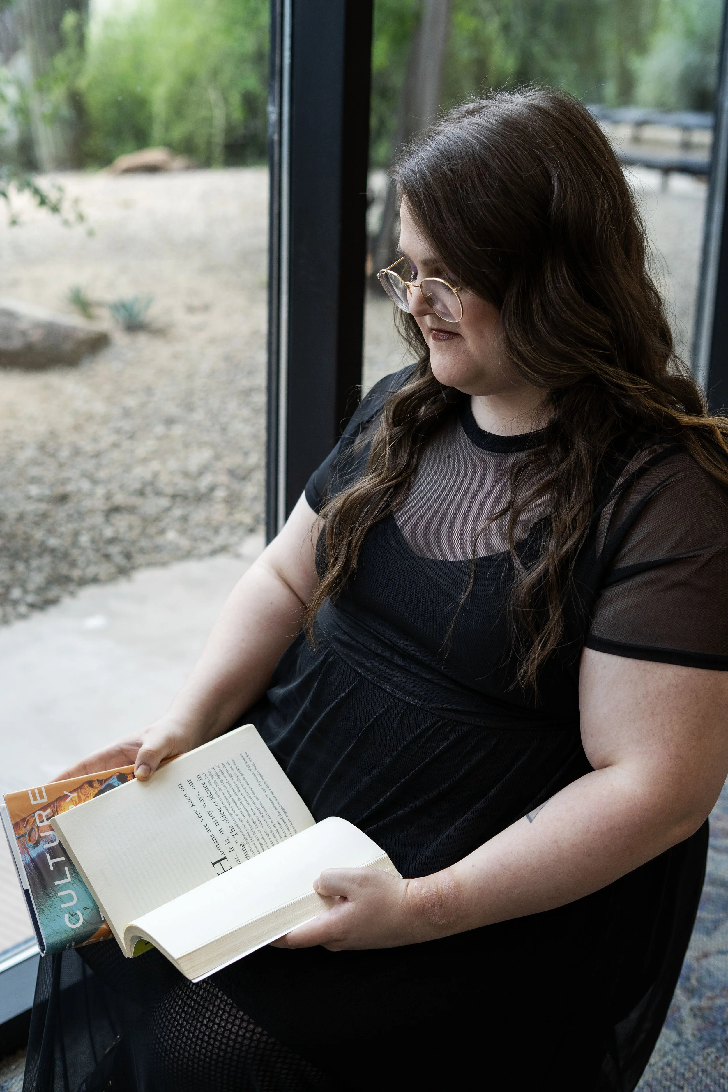 A woman with long wavy brown hair and glasses is reading a book while sitting by a large window, with trees and outdoor scenery visible outside.
