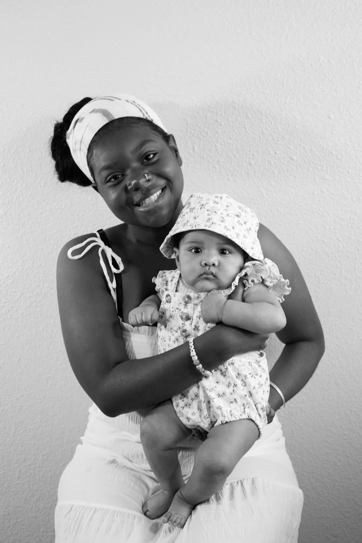 A smiling woman with a headscarf holding a baby girl in her arms, both facing the camera against a plain wall background.