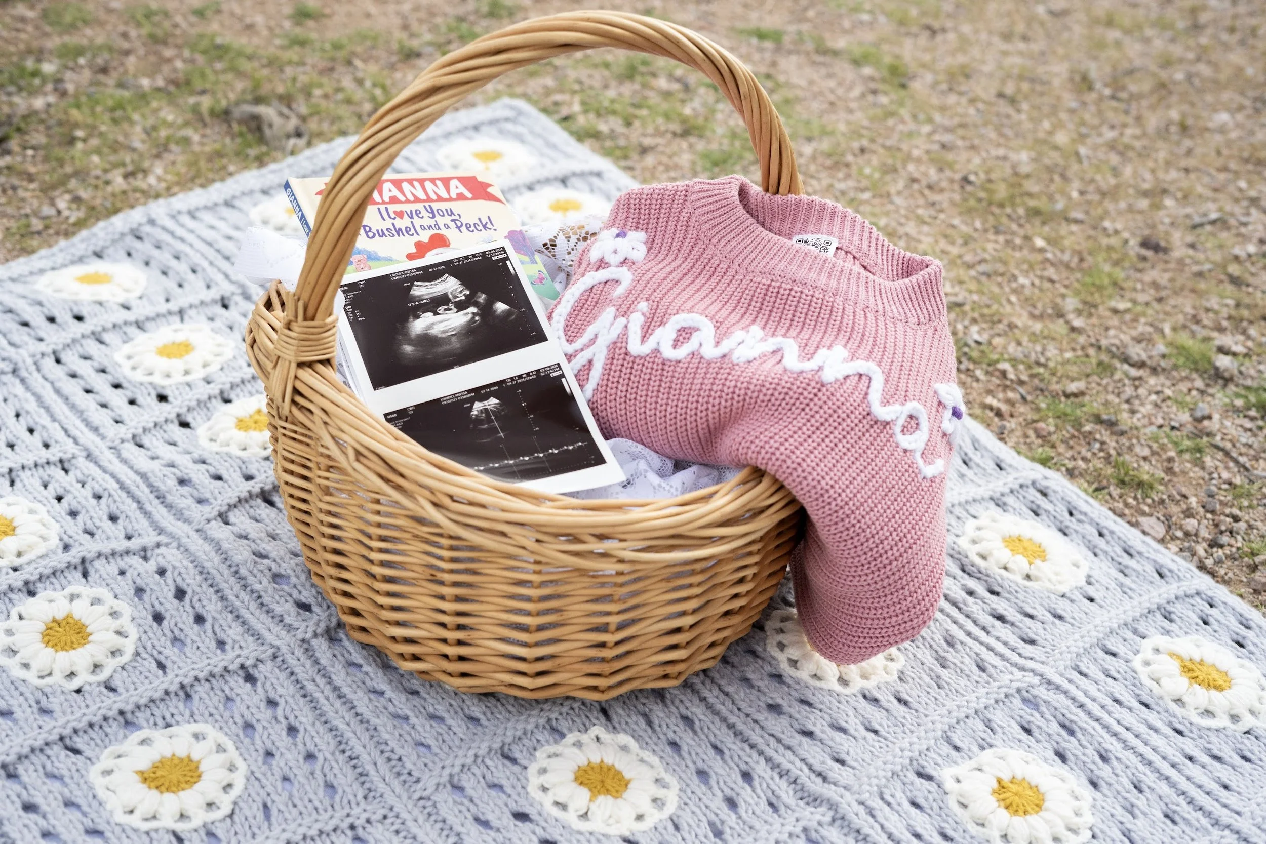 A picnic scene on a crocheted blanket with daisies, featuring a wicker basket containing ultrasound photos, a pink 'Glamour' sweater, and children's books, all outdoors on grass.