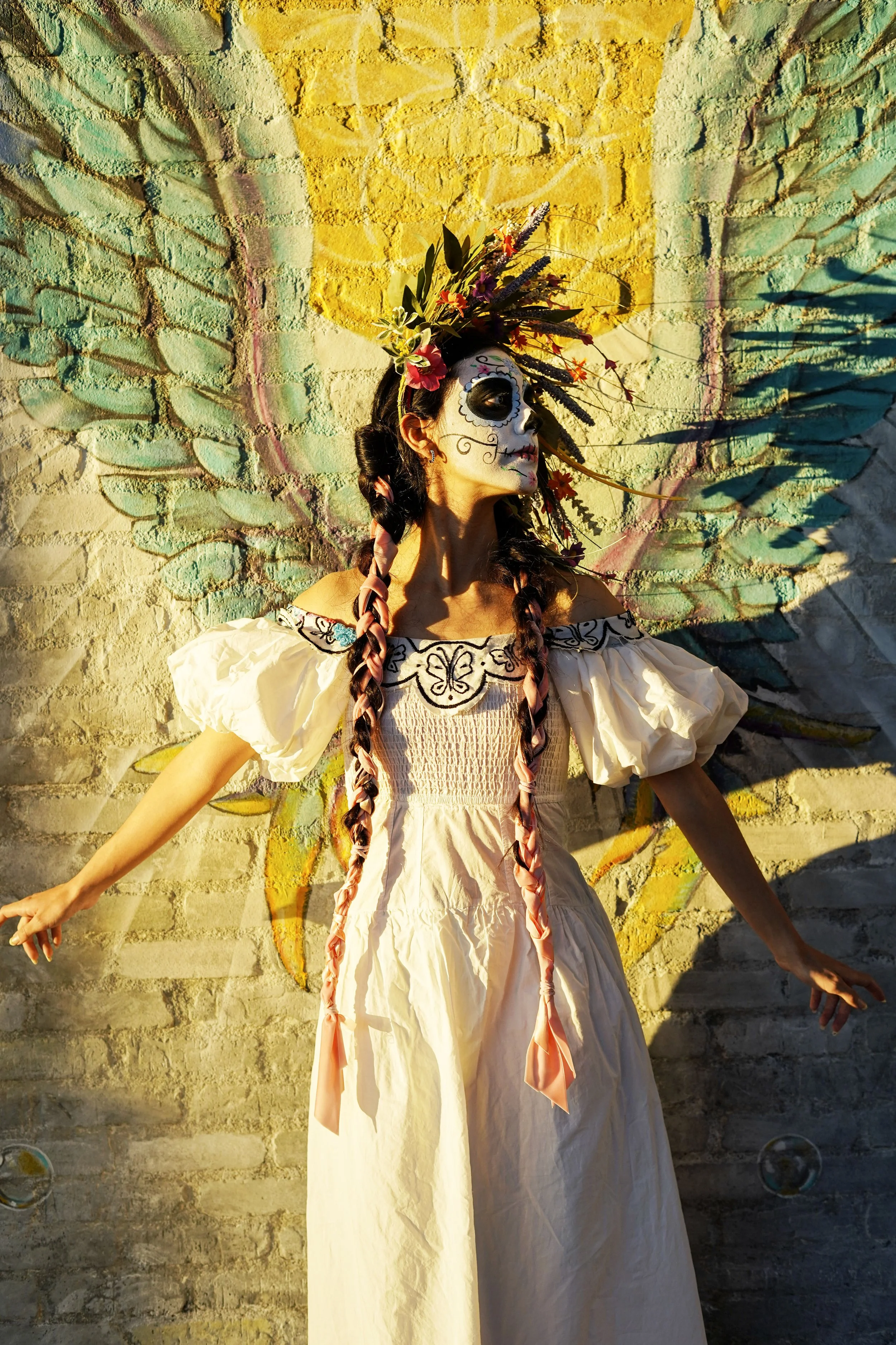 A woman dressed as a Day of the Dead calavera with face paint, a flower crown, and a long white dress, standing with arms outstretched against a wall with colorful wing artwork.