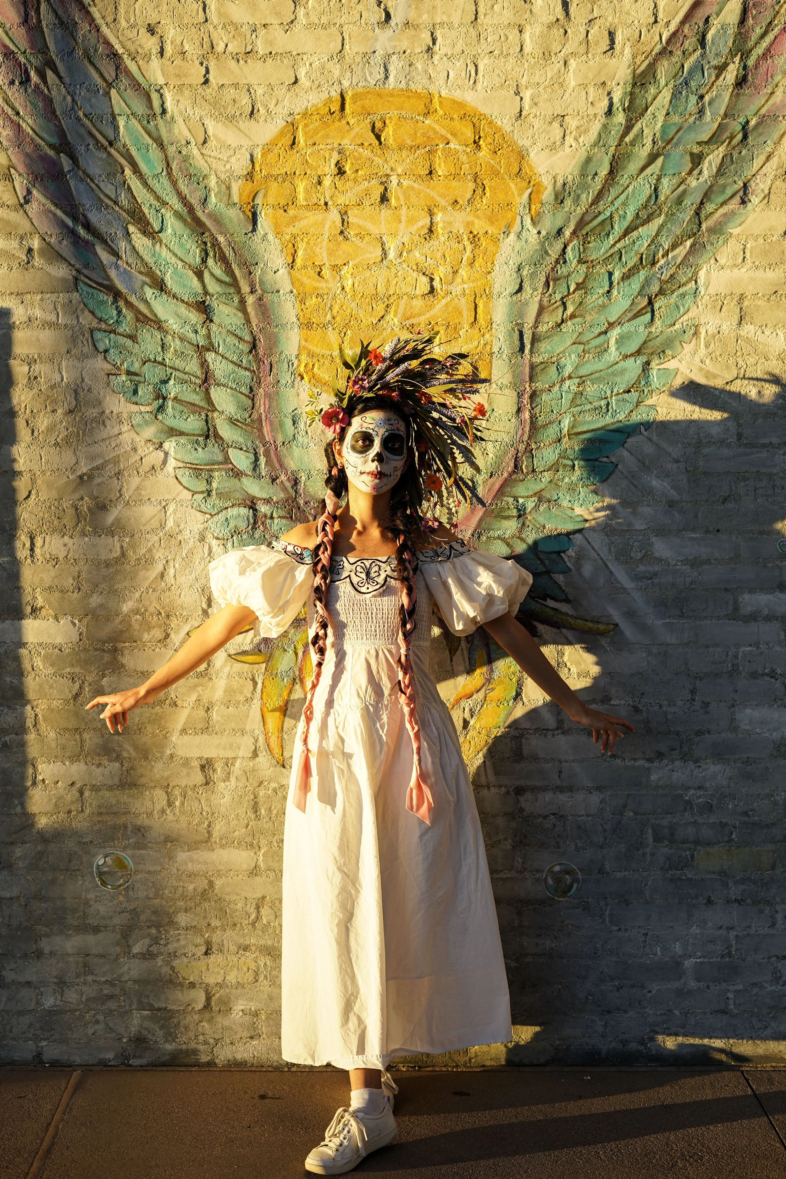 A person dressed as a sugar skull with face paint, wearing a white dress, standing in front of a mural of a colorful angel with large wings painted on a brick wall.