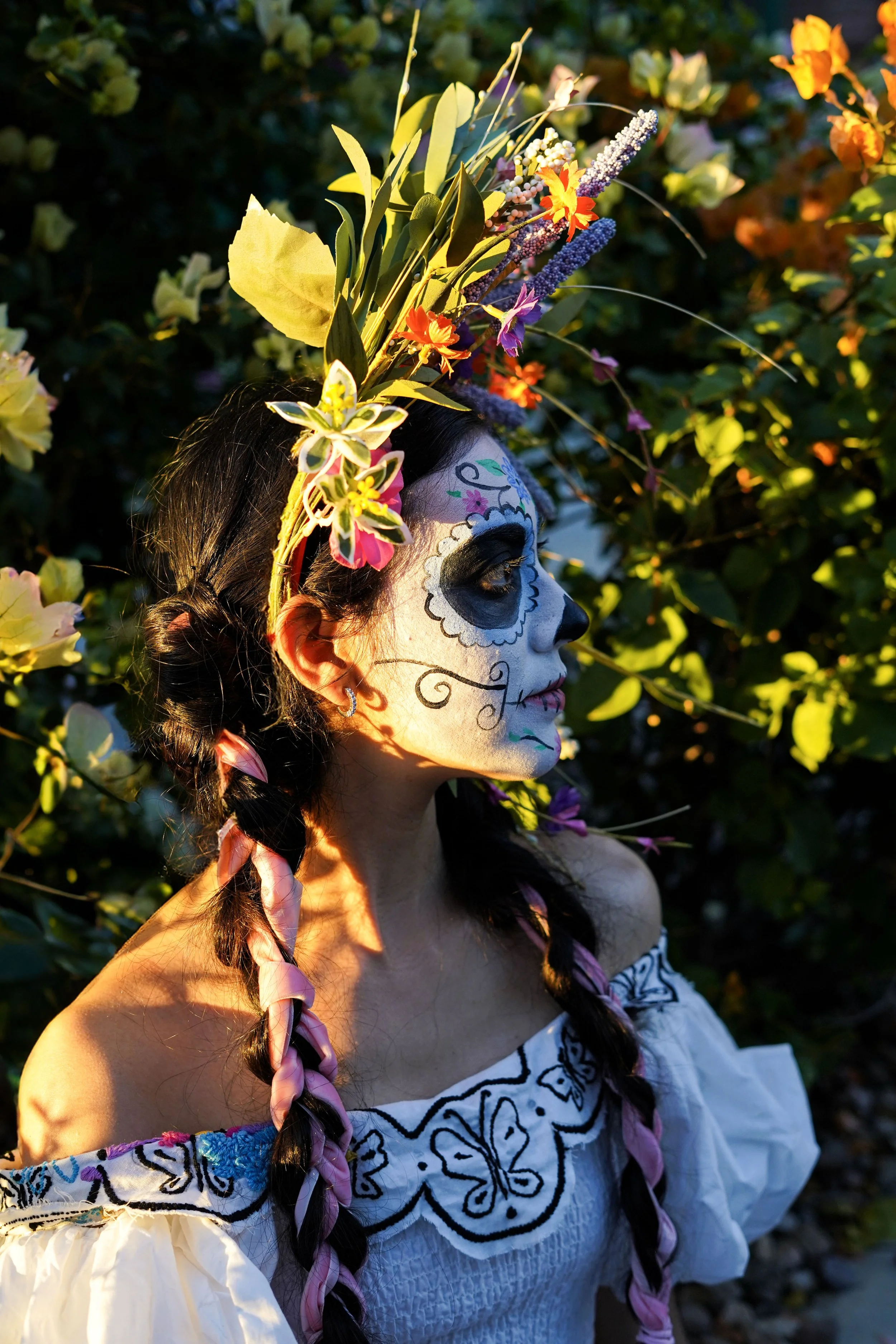 A woman in traditional Mexican attire with Day of the Dead face paint, wearing a floral crown, and standing among flowering bushes in sunlight.