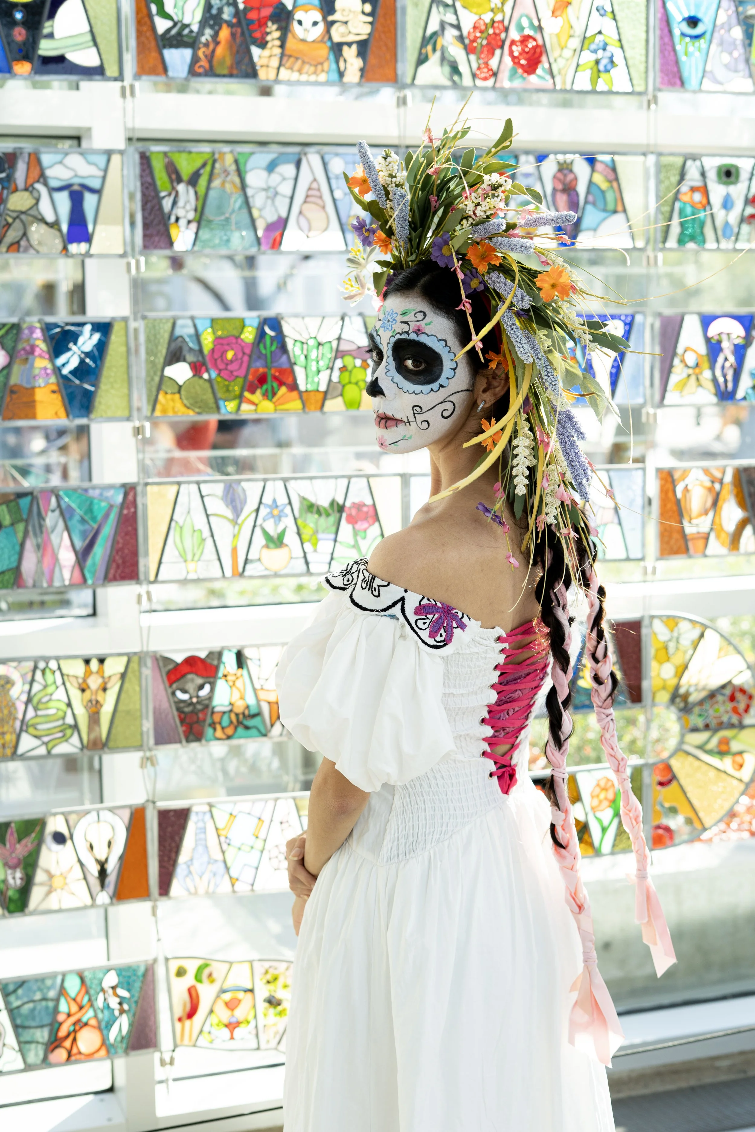 Woman dressed in a white dress with colorful embroidery and pink lace-up on the back, wearing traditional Mexican Day of the Dead face paint, with a large floral headdress, standing in front of a stained glass window with various colorful designs.