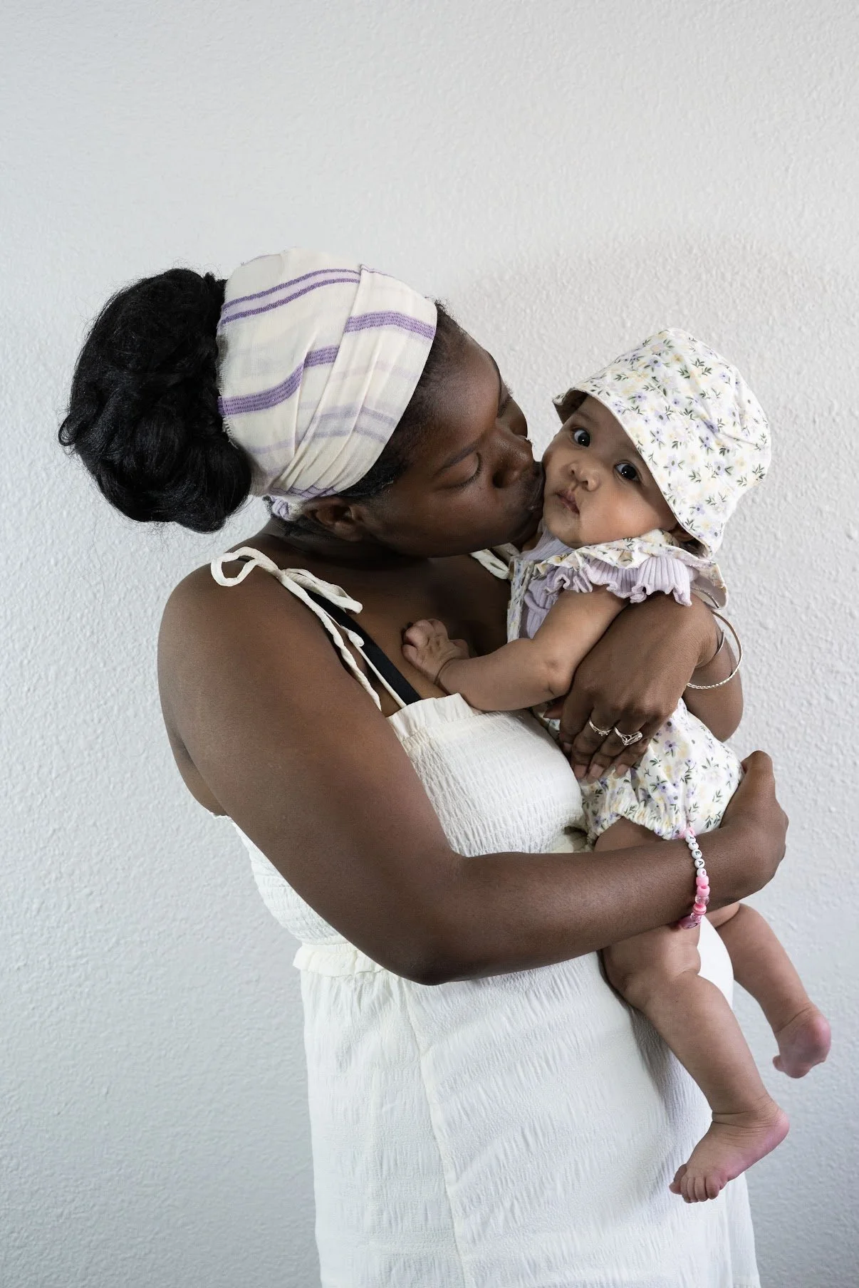 A woman wearing a white dress and a headscarf holding a baby girl hugging her, with the woman kissing the baby's cheek against a plain wall background.