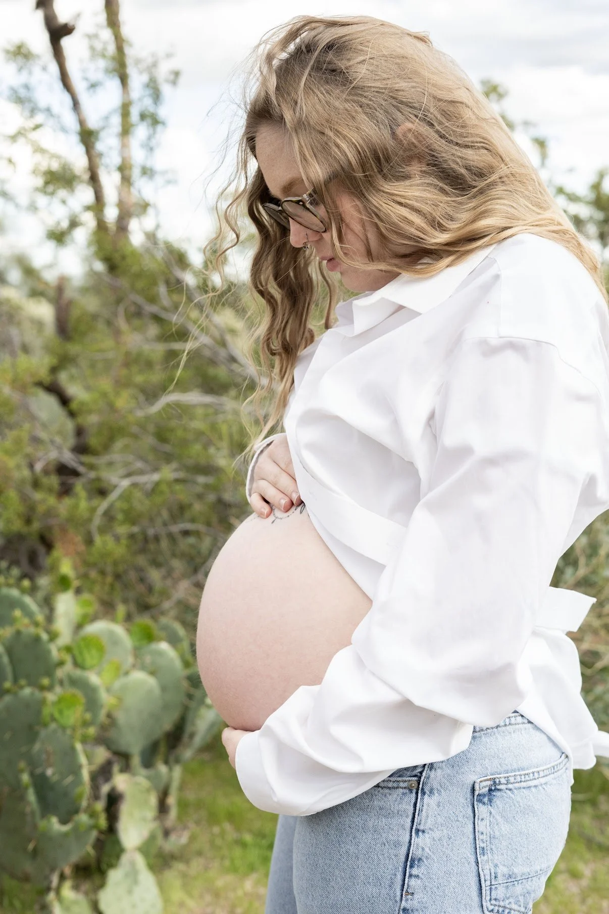 Pregnant woman in white shirt and jeans standing outdoors with cacti and trees in background, gently touching her belly.