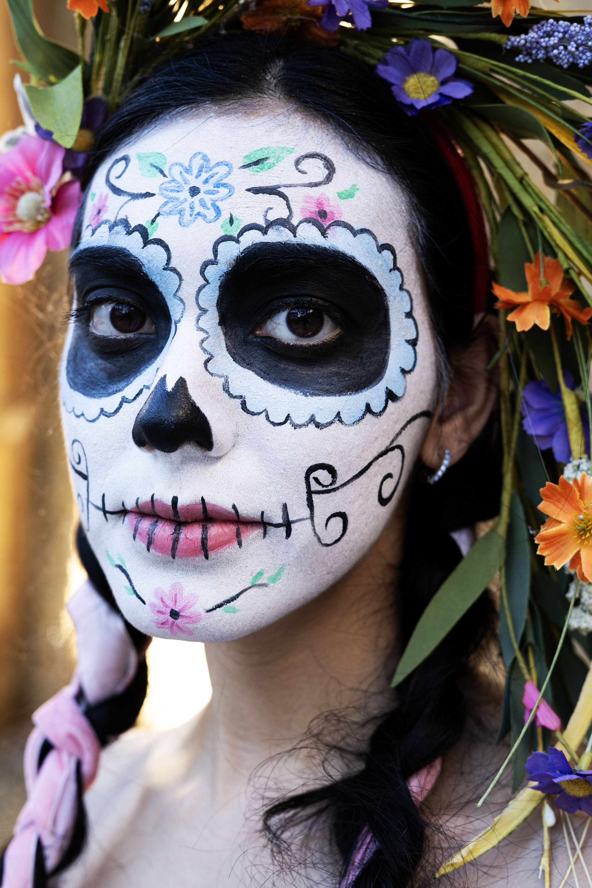 Person with face painted as a sugar skull, wearing a floral headpiece with colorful flowers and greenery.