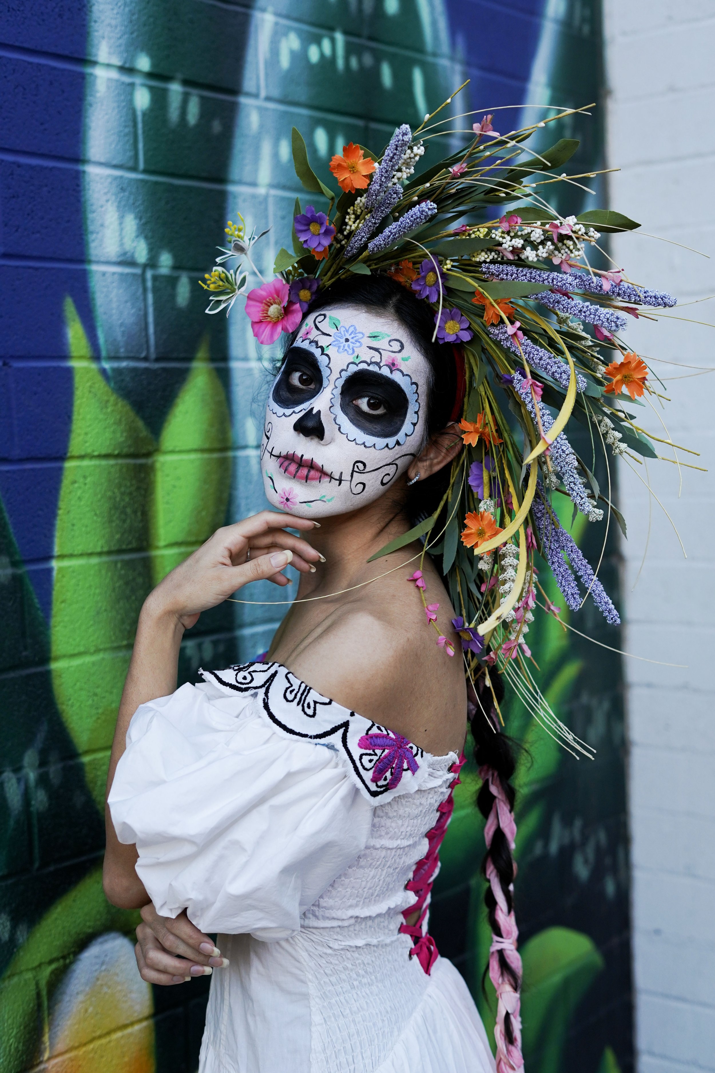 Woman with sugar skull makeup, floral headdress, and white off-shoulder dress standing against a colorful graffiti wall.