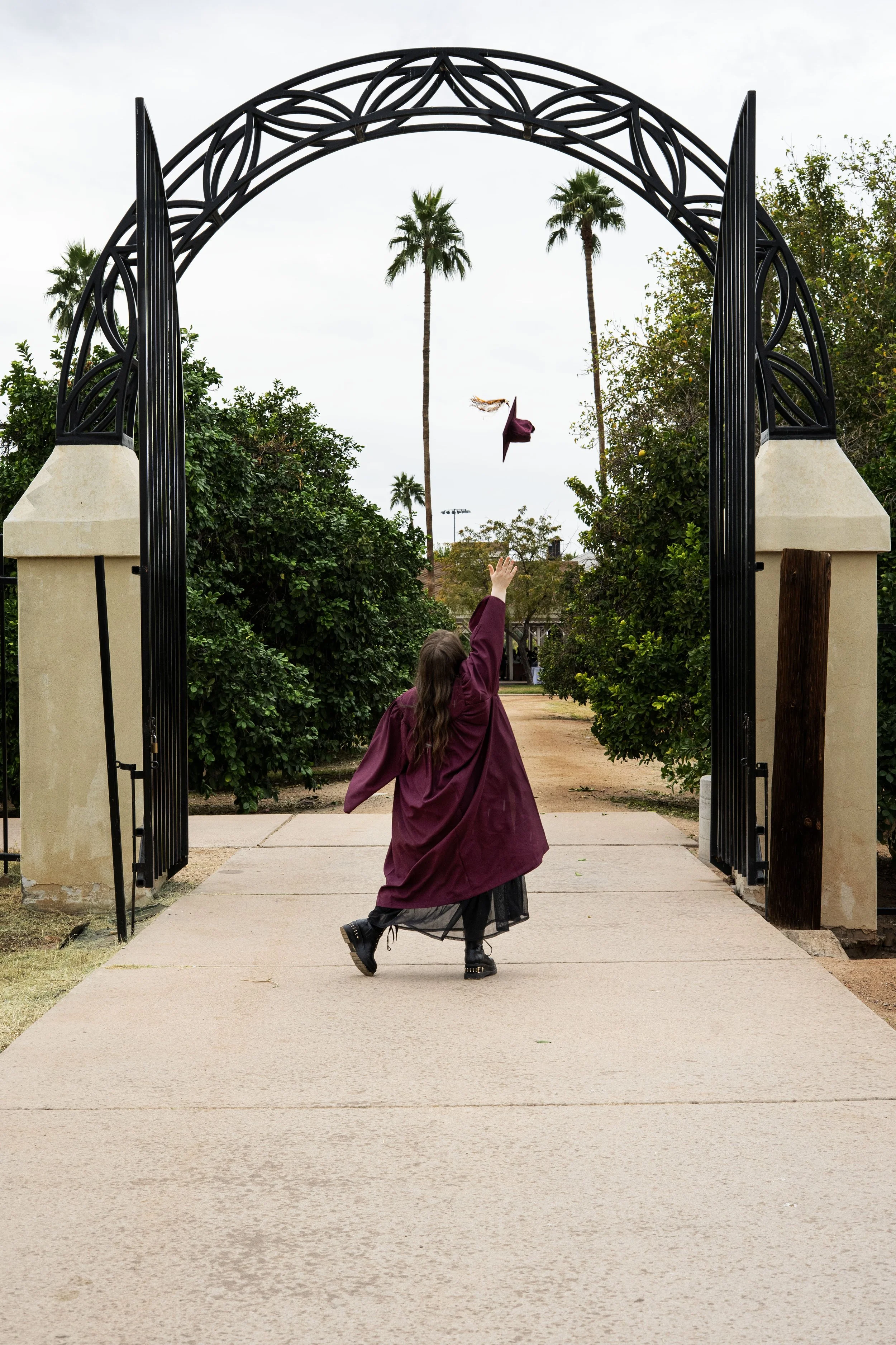 Woman in a purple graduation gown throwing her cap in the air at an outdoor venue with trees and palm trees in the background.