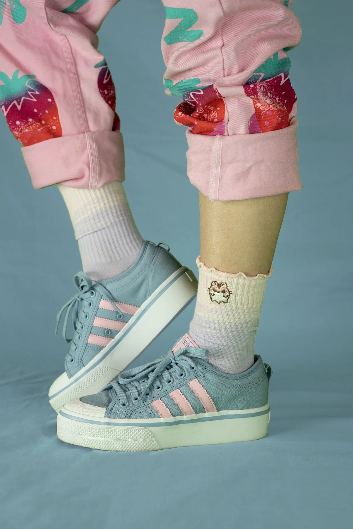 Close-up of a person wearing light blue Adidas sneakers with pink stripes, white socks with pink ruffles, and pink pants with strawberry and alphabet prints, against a blue background.