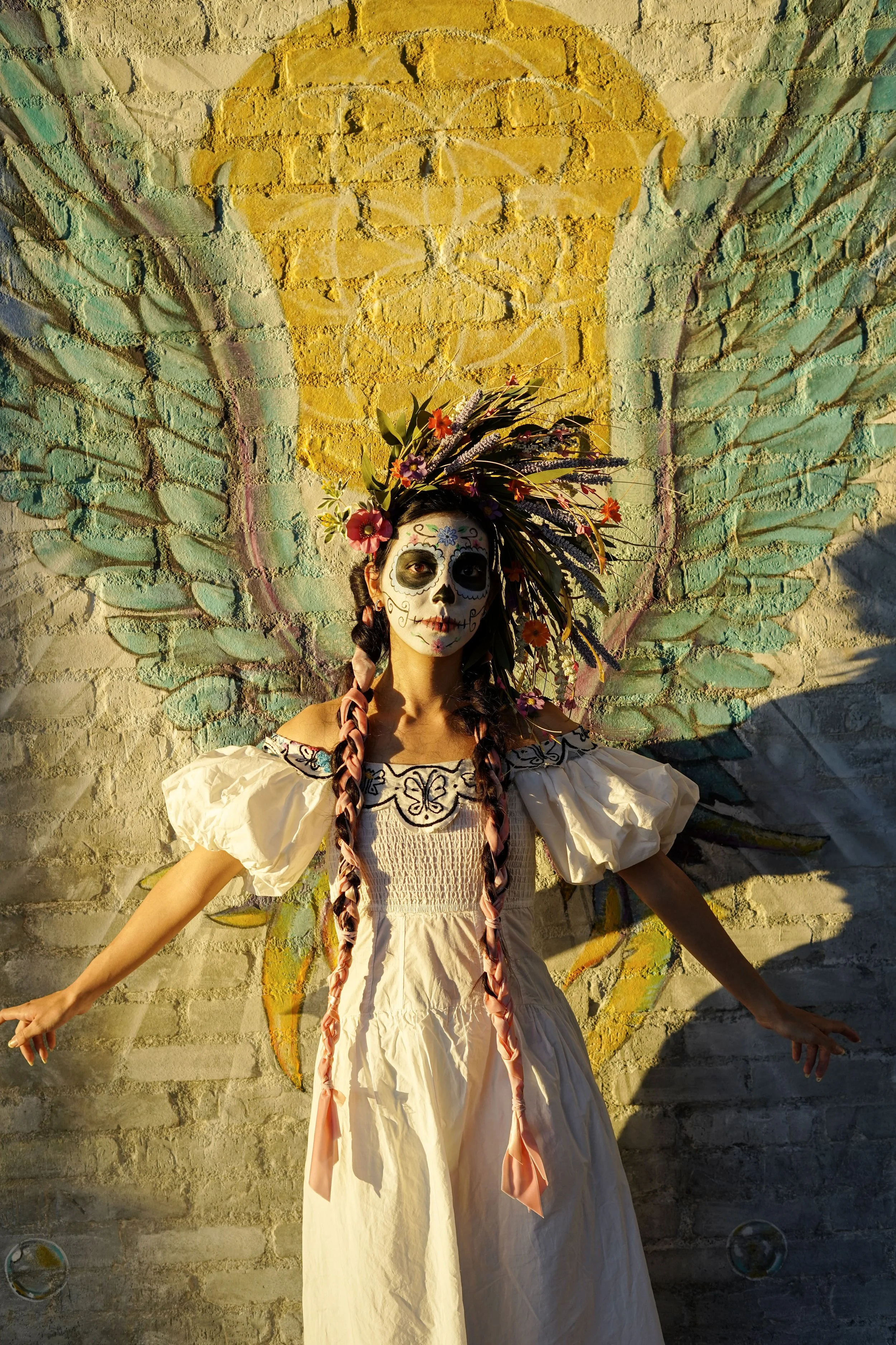 A woman dressed as a sugar skull with face paint, wearing a white dress with puffed sleeves and embroidered details, standing in front of a brick wall mural of angel wings with a yellow halo. She has long, braided hair with pink ribbons and a colorfu