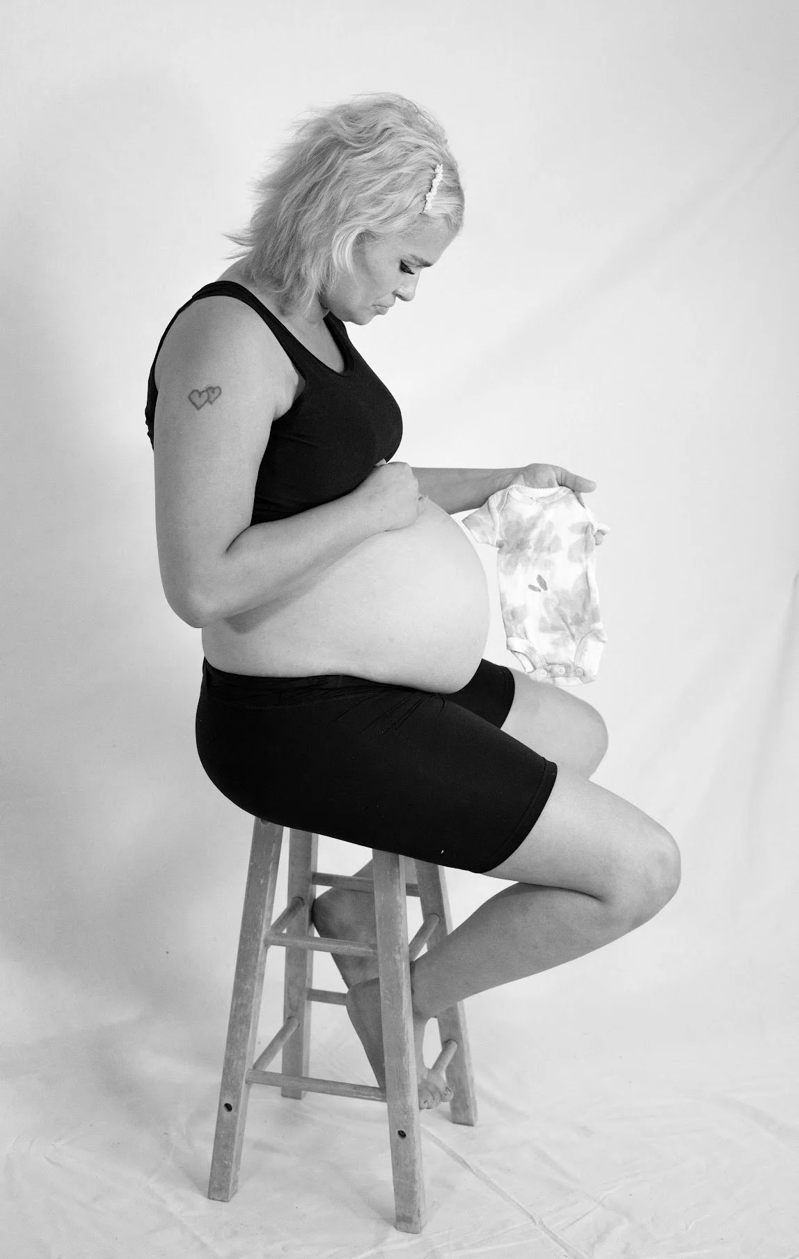 Pregnant woman sitting on a stool, holding a baby onesie, in a studio with a plain background.