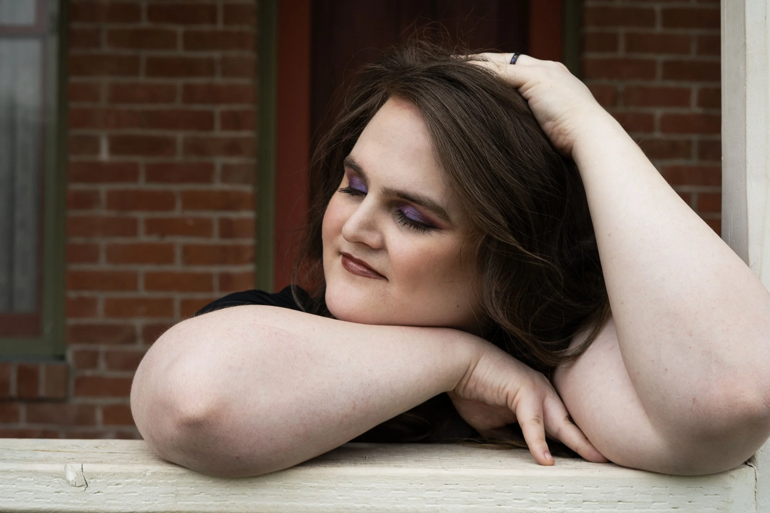 A woman with long brown hair and purple makeup resting her head on her arm with her eyes closed, leaning on a wooden surface outdoors with a brick wall in the background.