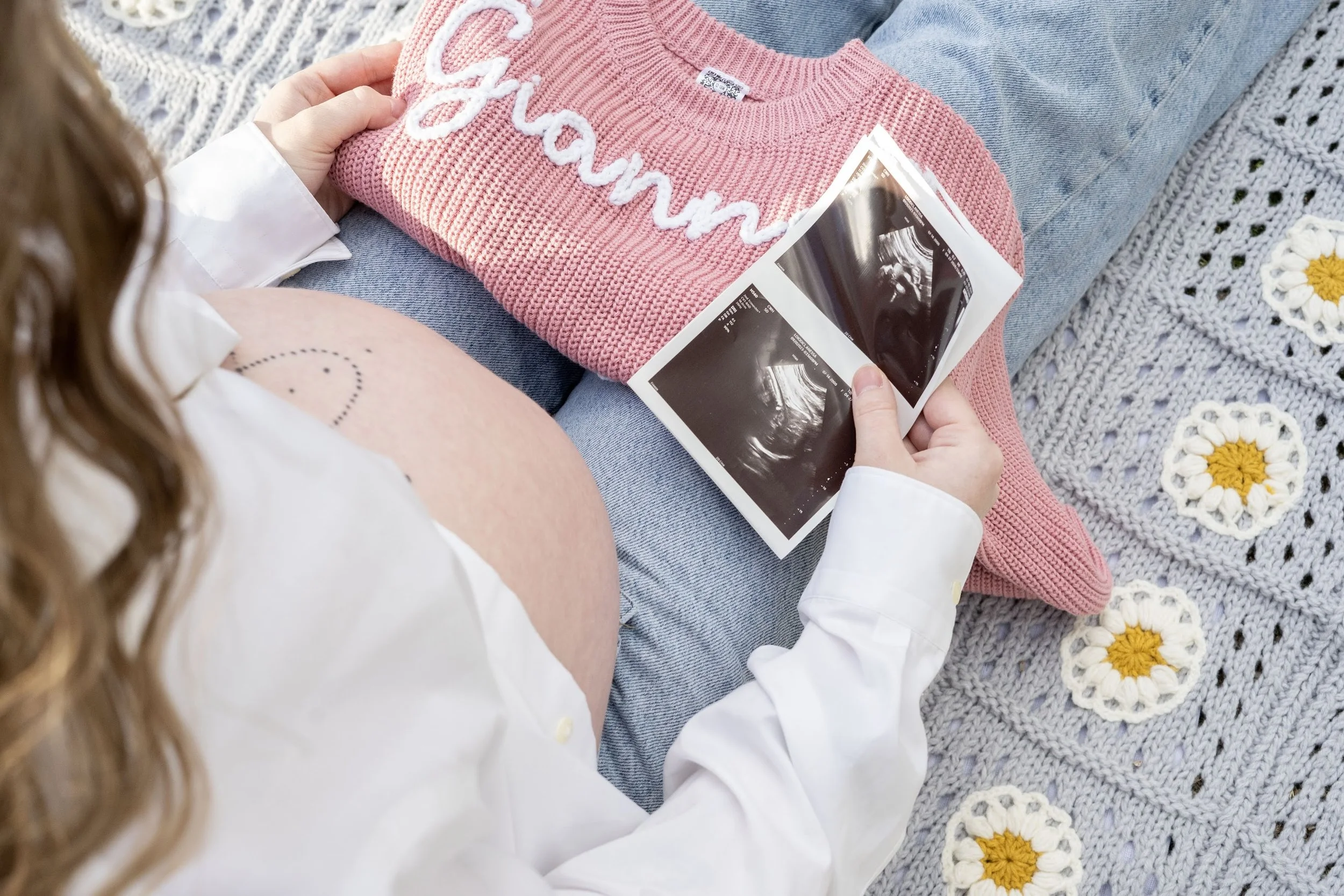 A pregnant woman sitting on a knitted blanket with daisies, holding ultrasound images, wearing a white shirt, has a tattoo on her arm and a pink sweater with white lettering on her lap.