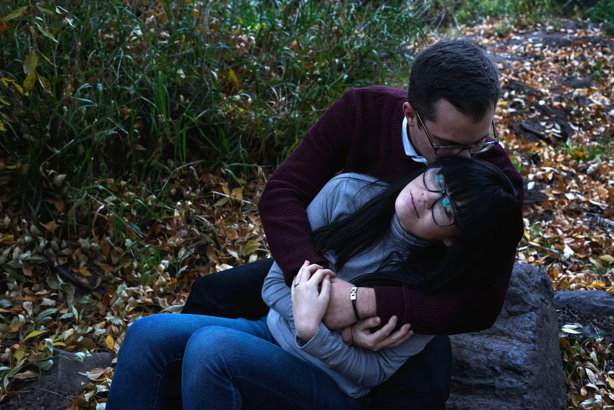 A man is comforting a woman who appears to be upset, sitting on a rock in a wooded area with fallen leaves.