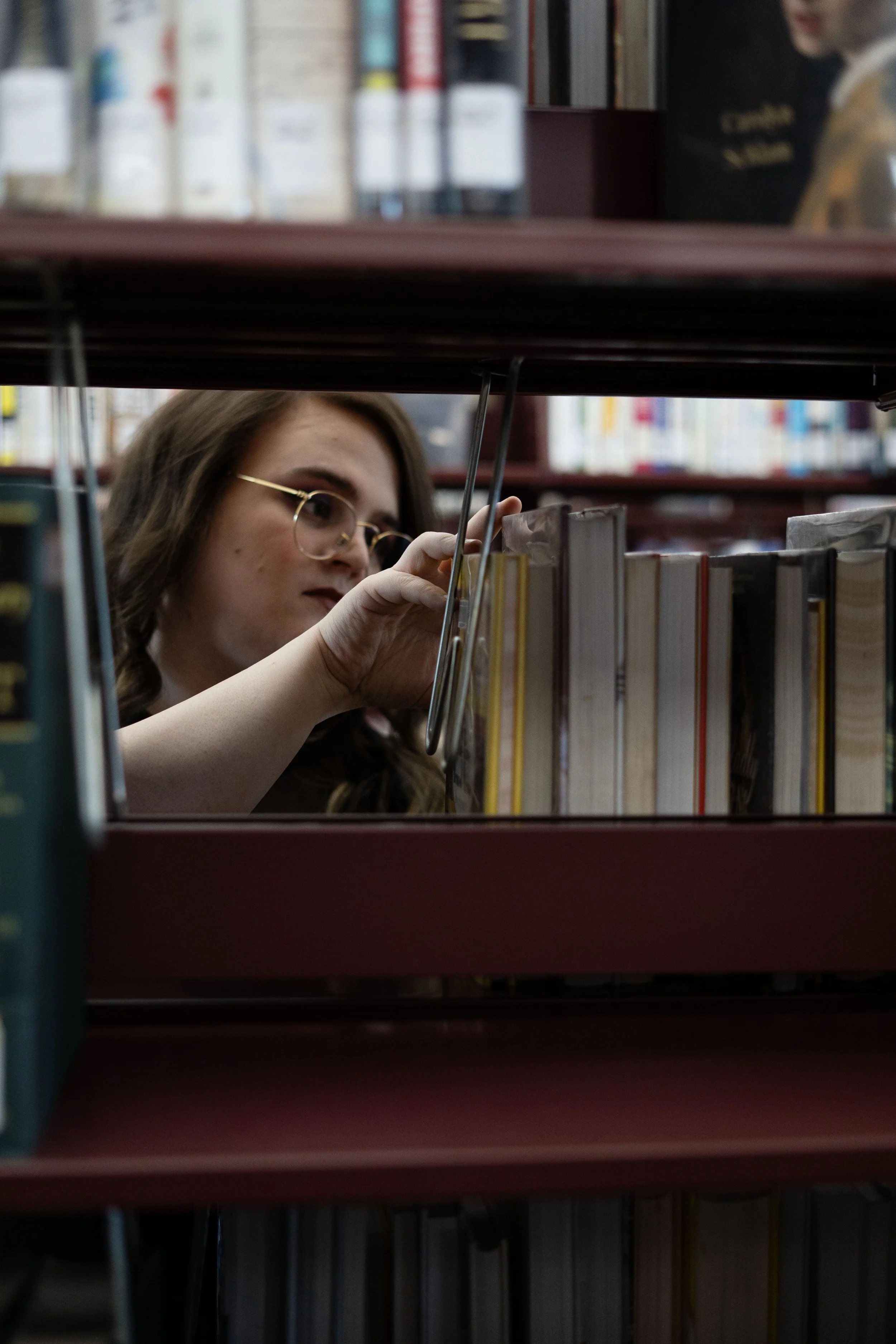 A woman with glasses browsing books on a library shelf, viewed through a gap between shelves.