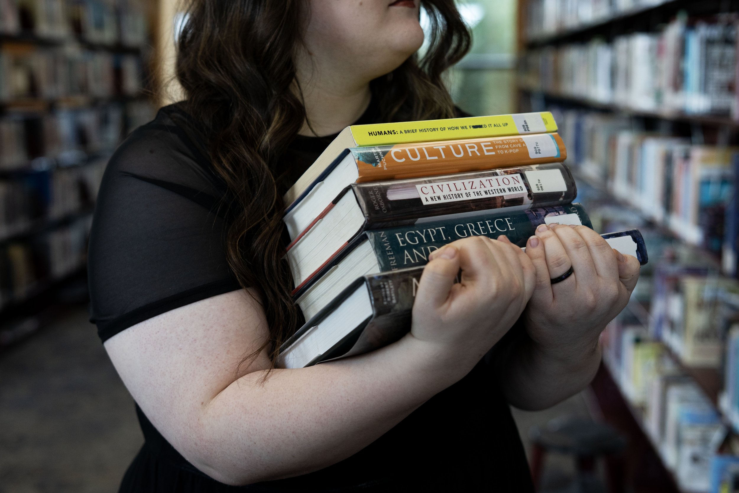 Person carrying multiple books in a bookstore or library, with shelves filled with books in the background.
