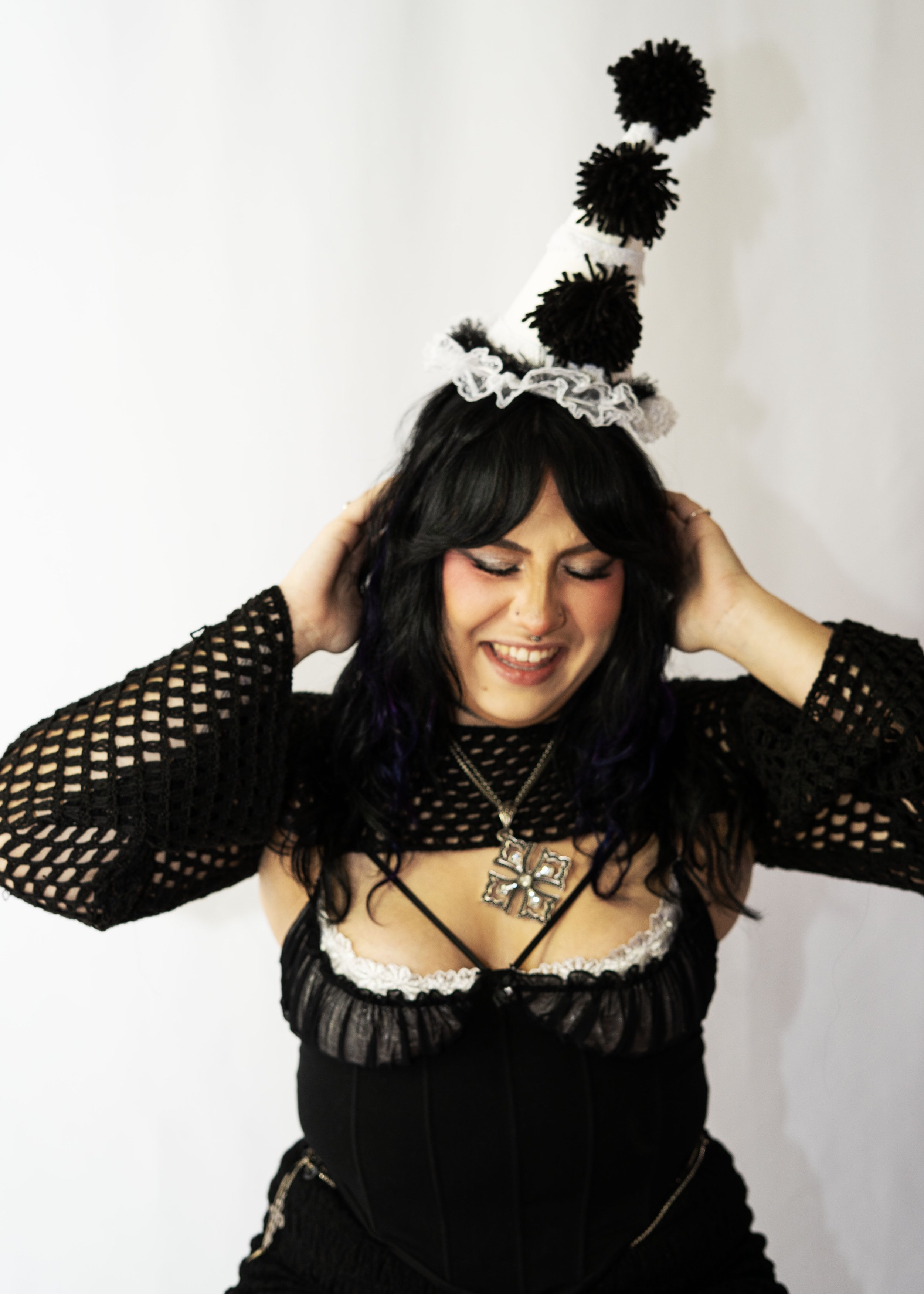 A woman with dark hair and makeup, wearing a black mesh top and a necklace with a large cross, is adjusting a whimsical white and black striped party hat with pom-poms on her head, while smiling with her eyes closed.