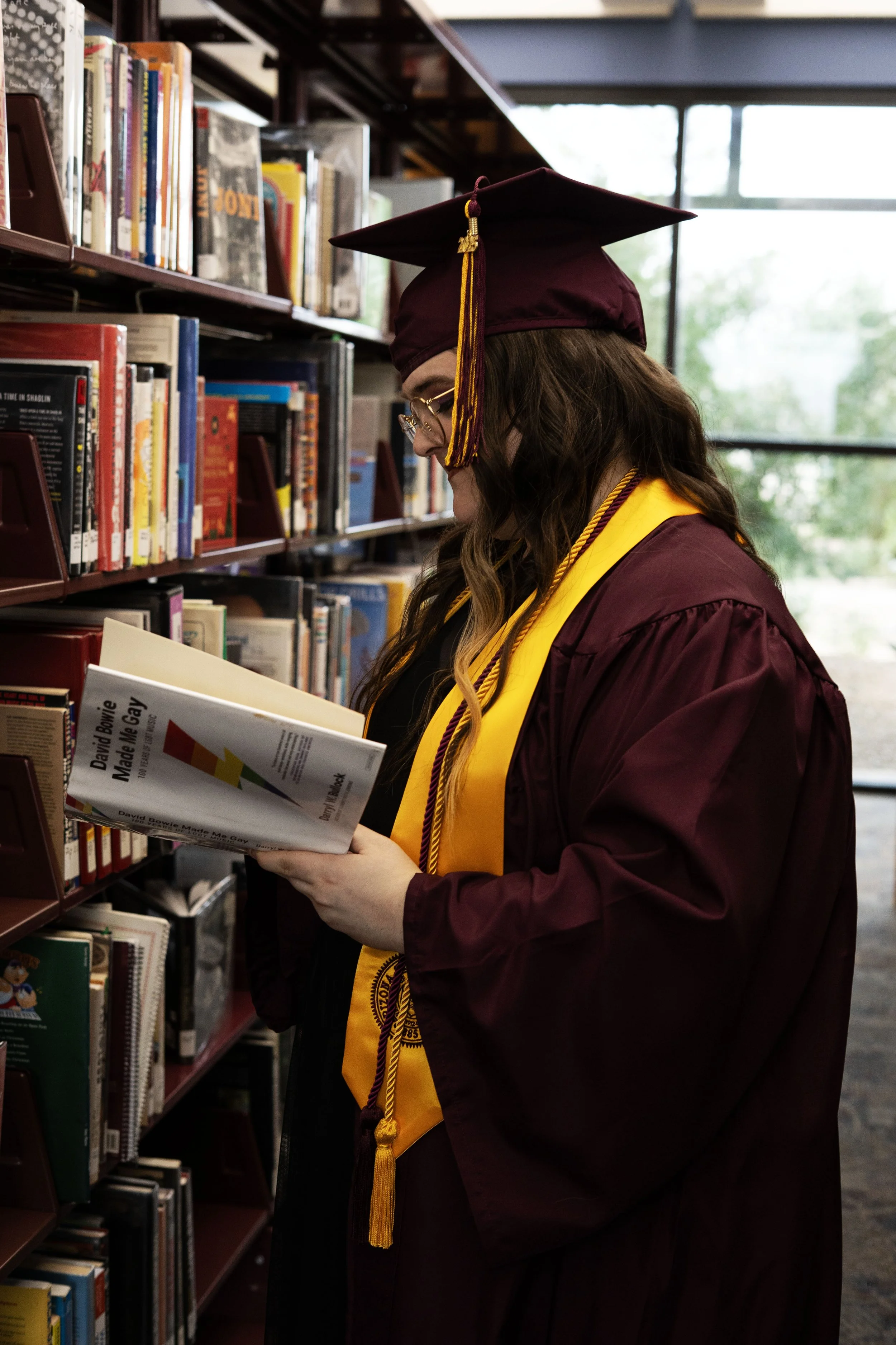 A woman dressed in maroon graduation gown and cap with a yellow stole and tassel, reading a book in a library surrounded by bookshelves.