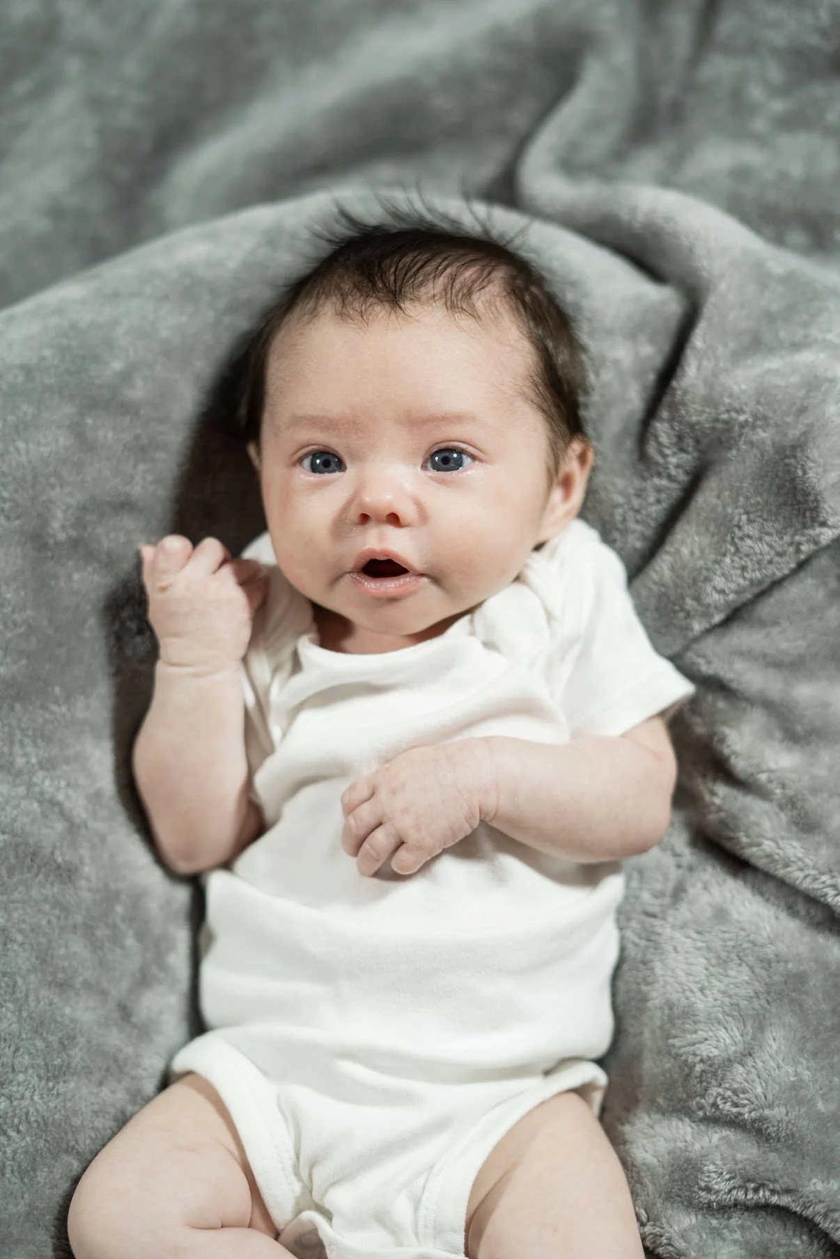 A baby with blue eyes lying on a gray blanket, wearing a white onesie.