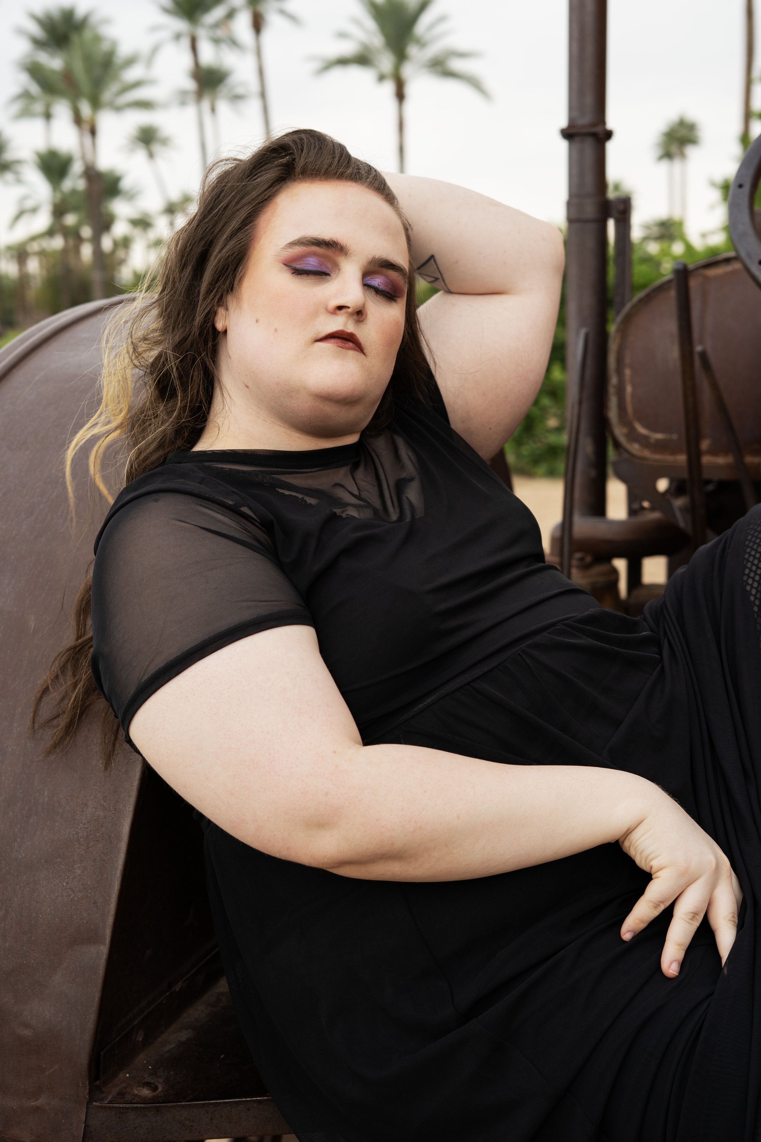 A woman with long brown hair, purple eye makeup, and dark lipstick, wearing a black mesh top, relaxing outdoors against a vintage vehicle, with palm trees in the background.