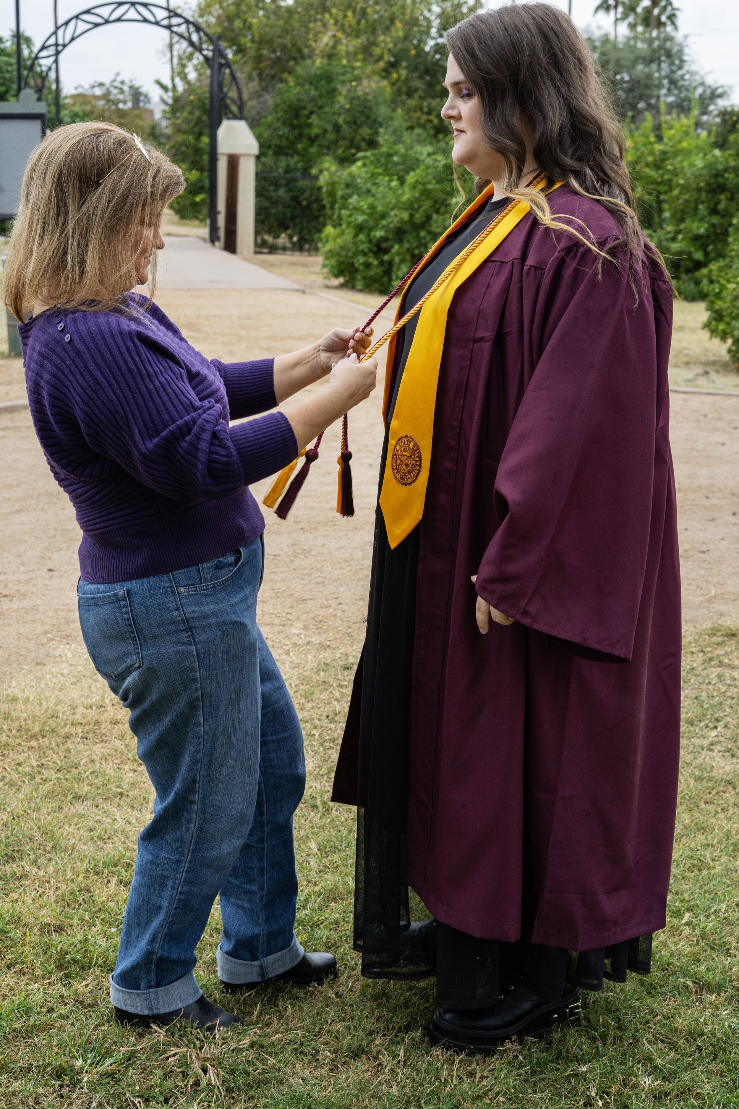 A young girl in a purple sweater is helping a woman in a maroon graduation gown and a yellow honor cord.