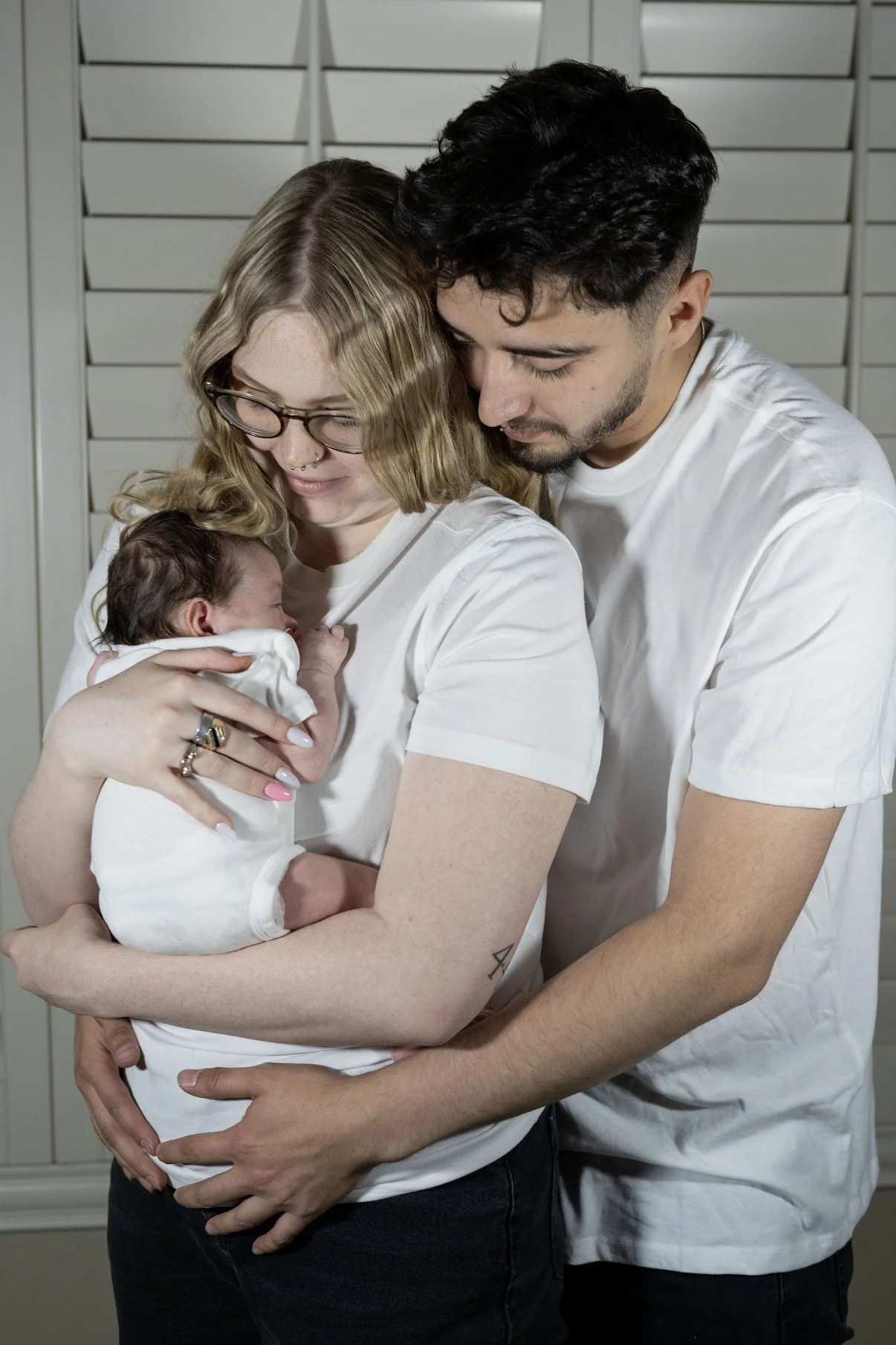 A woman holding a newborn baby, with a man embracing both of them, all dressed in white shirts, in a warm family moment.