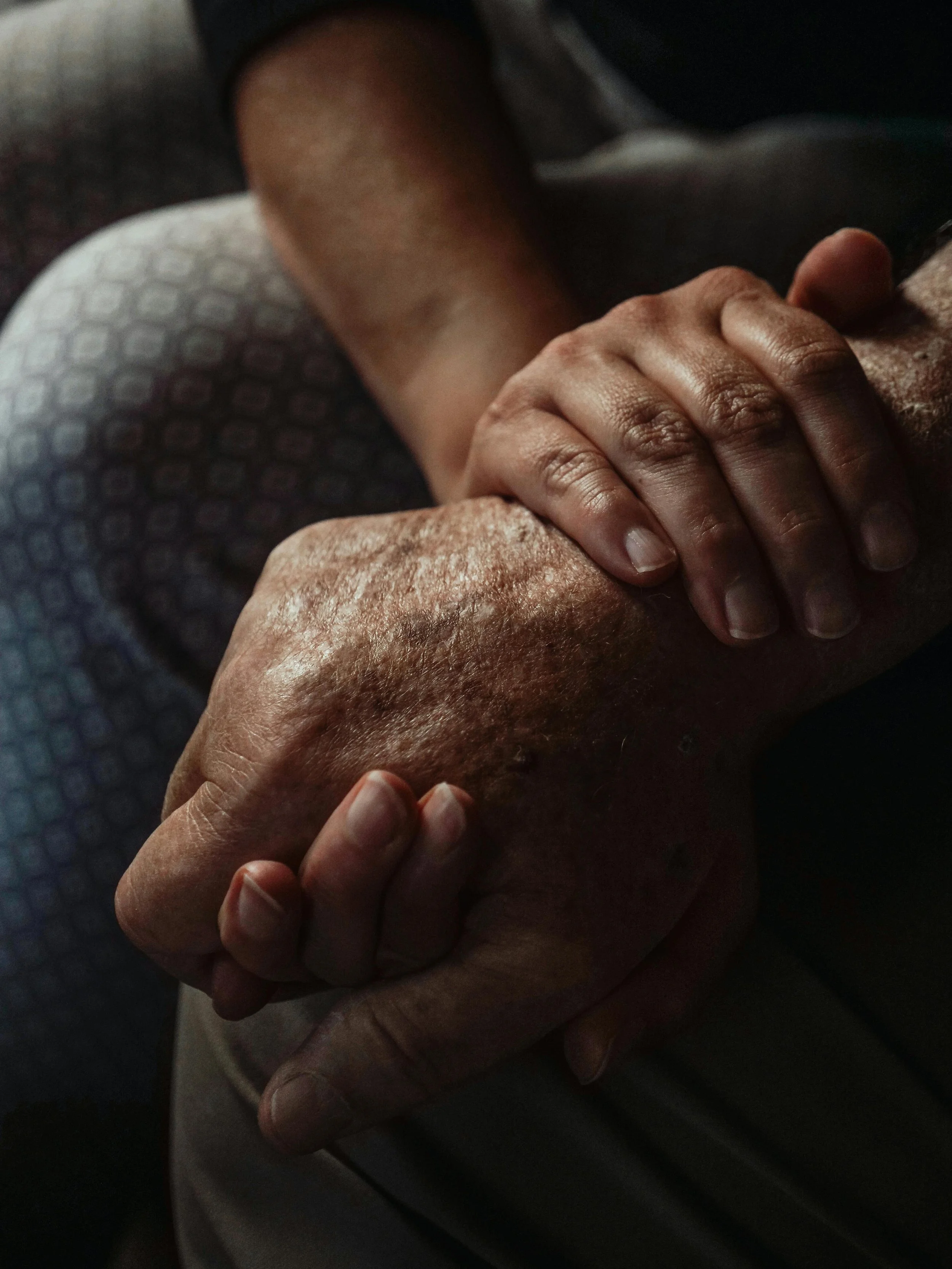 Close-up of two hands holding each other, one hand over the other, with the background showing part of a person's body.