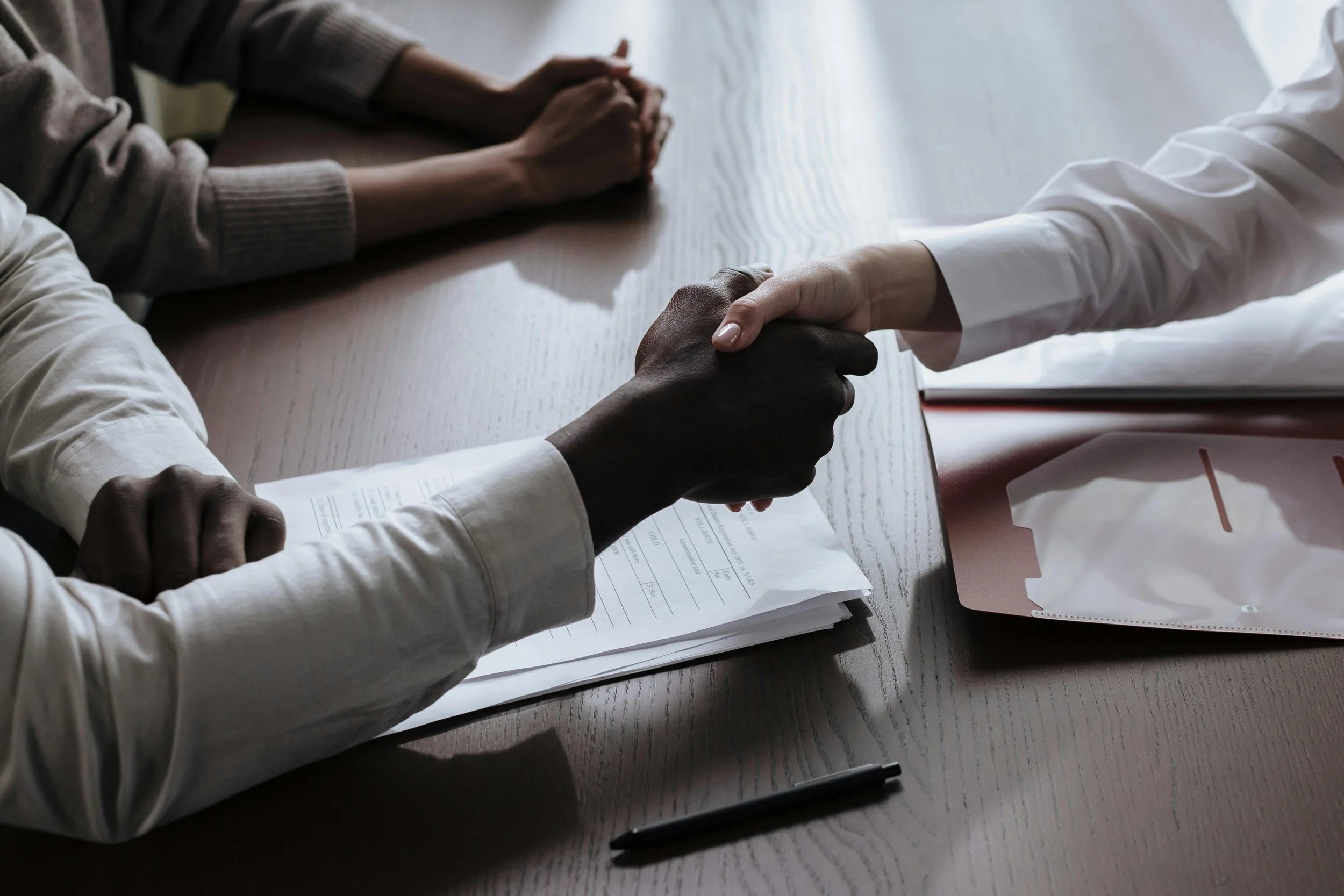 Two people holding hands during a meeting. One person has darker skin and the other has lighter skin, both wearing professional attire. Papers, notebooks, and pens are on the table.