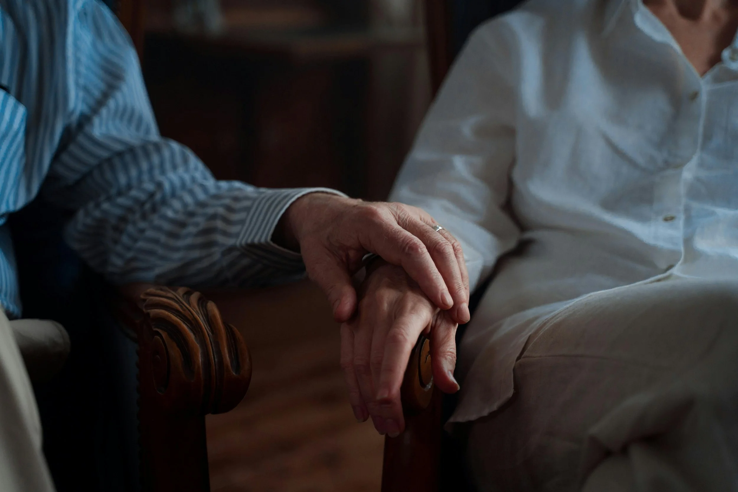 Two elderly people holding hands, sitting together, wearing casual clothing, in a cozy indoor setting.
