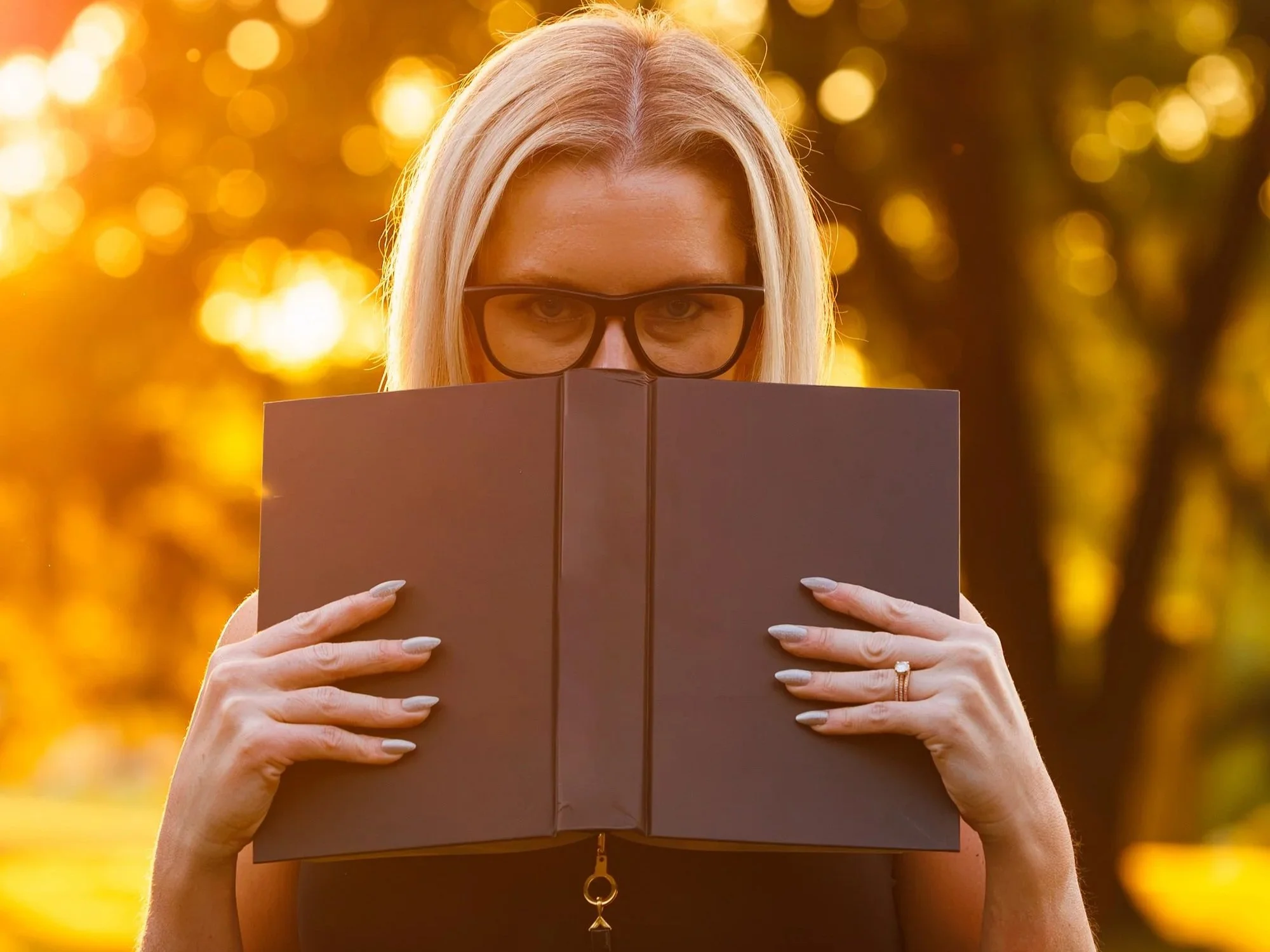 A woman with blonde hair and glasses holding a dark-colored book in front of her face, outdoors during sunset with a blurred background of trees and golden light.