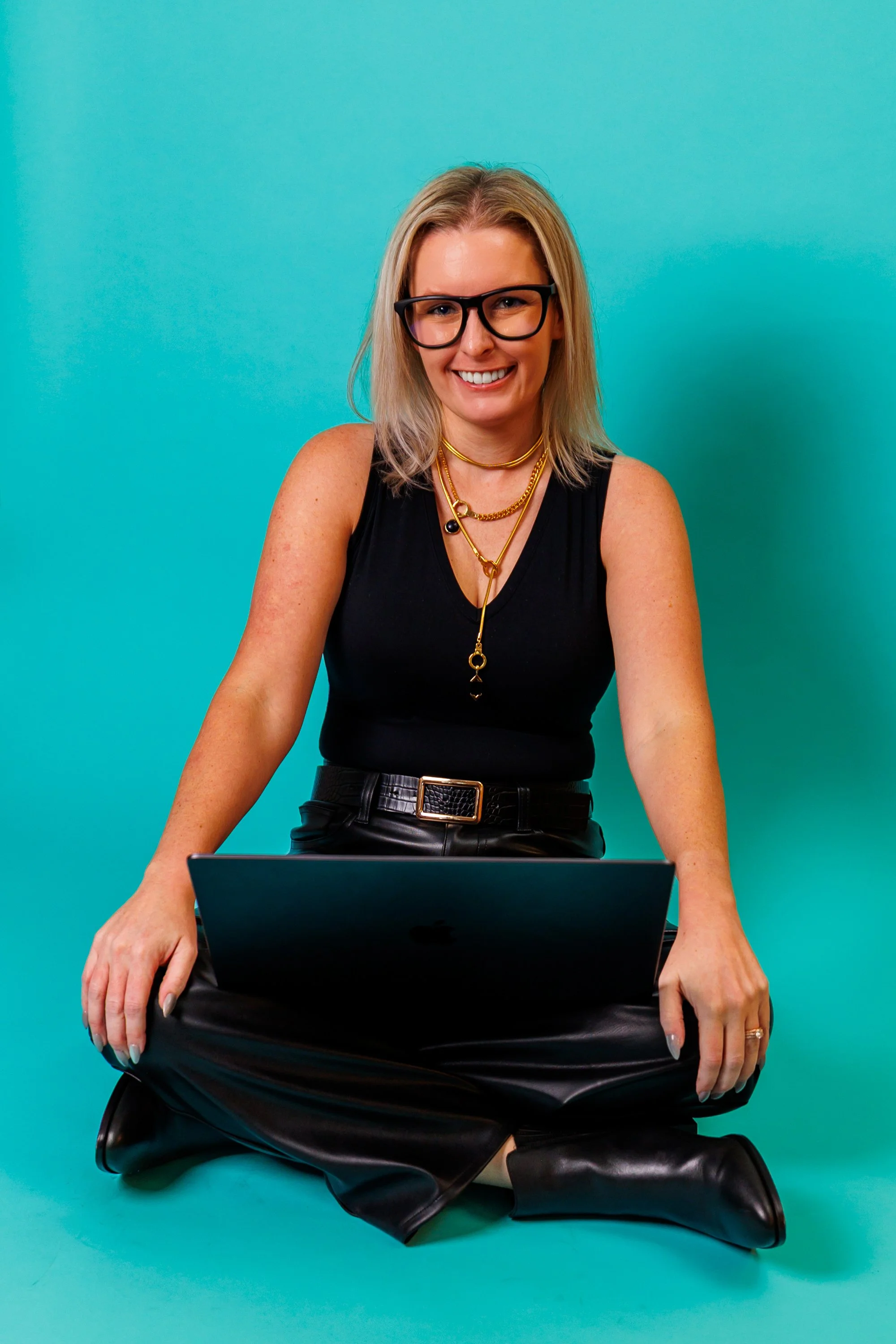 A woman with blonde hair, wearing black glasses, a black sleeveless top, and gold jewelry, is sitting cross-legged on the floor with a laptop on her lap against a blue background.