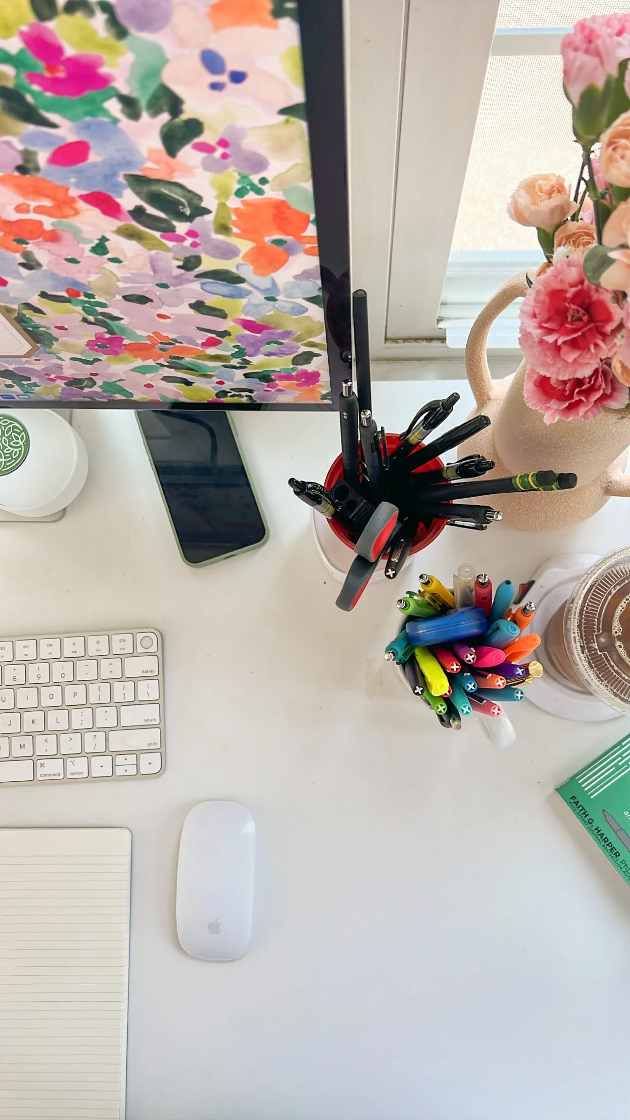 Desk with a colorful computer monitor, wireless keyboard and mouse, smartphone, two containers of multi-colored pens, a pink vase with pink flowers, and a hardcover book, near a window.