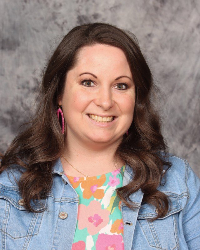 A smiling woman with long wavy brown hair, wearing pink hoop earrings, a colorful floral top, and a light blue denim jacket, against a gray textured background.