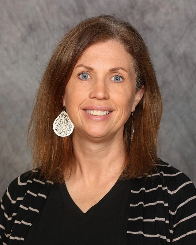 A woman with shoulder-length brown hair, blue eyes, smiling, wearing a black top with a black and white striped cardigan and large white earrings, against a gray textured background.