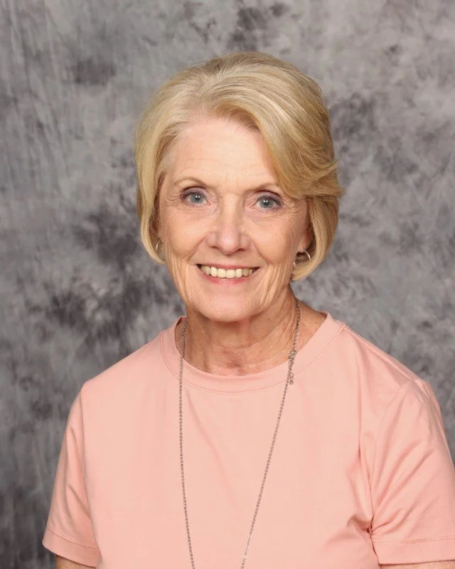 A woman with long blonde hair, smiling, wearing a black top, a light-colored scarf, and a black necklace with a round pendant, against a gray background.