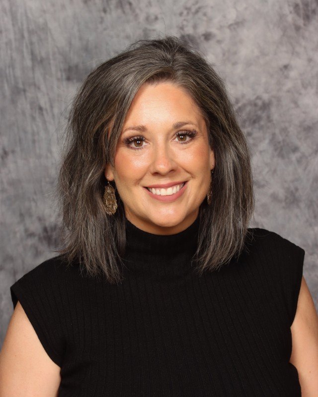 Close-up of a man with dark curly hair, wearing clear glasses, a nose ring, and a dark green sweater. He has light stubble and a thoughtful expression. In the background, part of a white wall and a green plant are visible.
