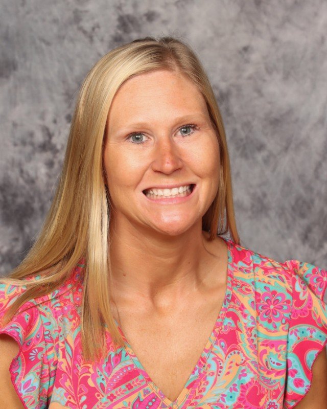 A woman with long blonde hair, smiling, wearing a colorful pink, blue, and yellow patterned blouse, against a gray mottled background.