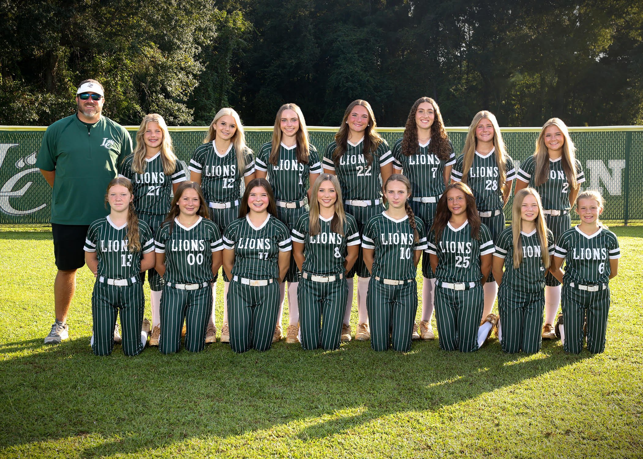 A girls' softball team poses for a team photo on a field, with 13 players and a coach, all wearing green striped uniforms with 'LIONS' written on the front, in front of a fence with trees in the background.