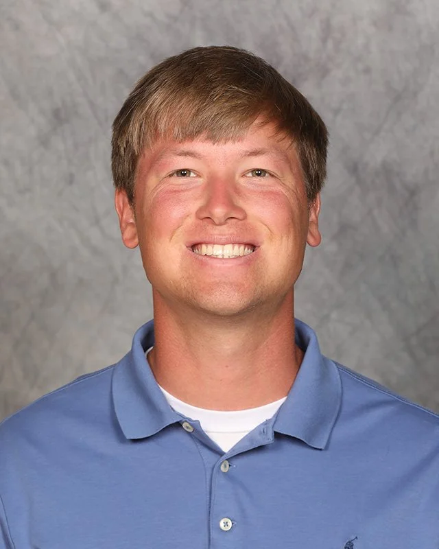 Headshot of a smiling man with short light brown hair, wearing a blue collared polo shirt against a neutral gray background.