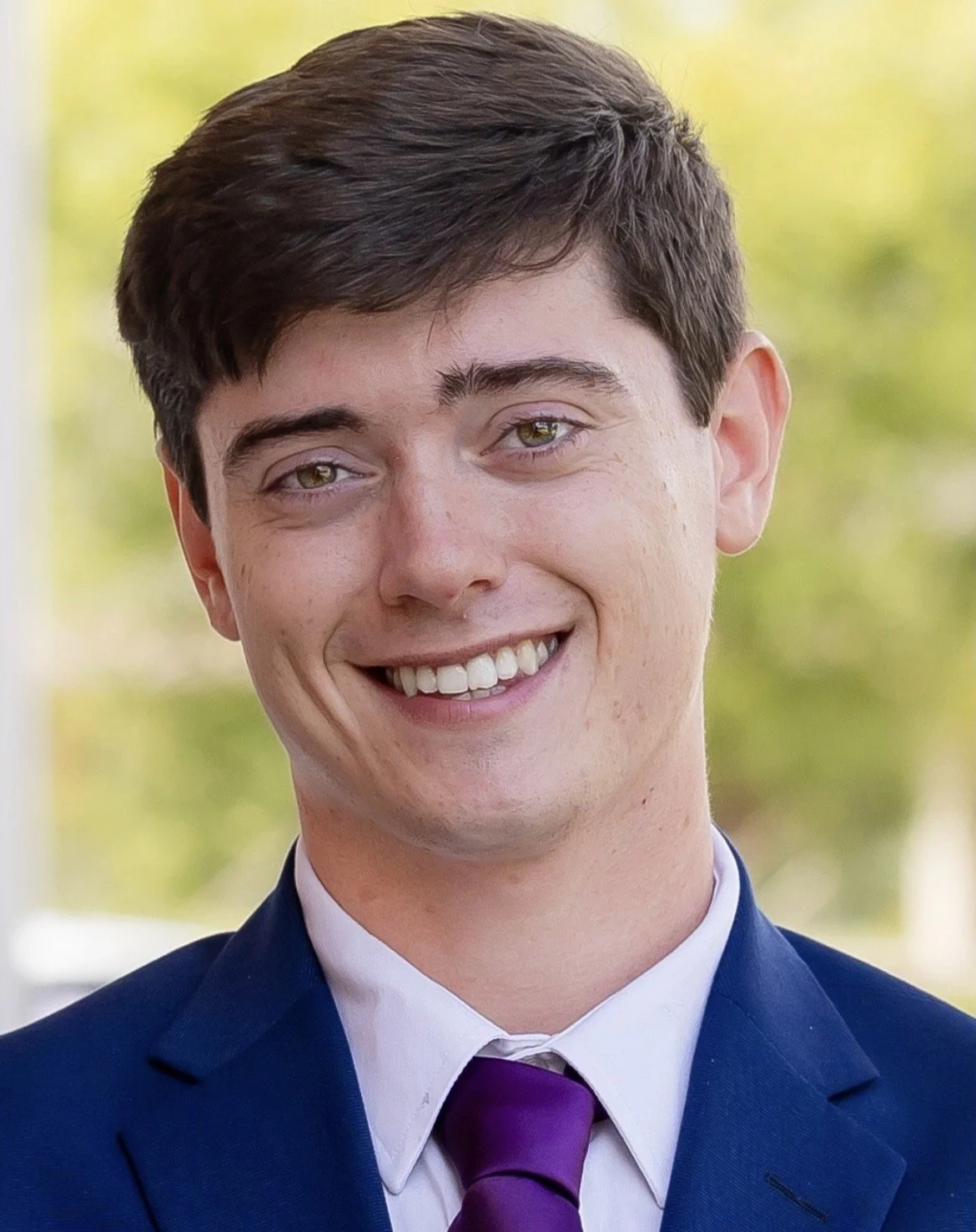 Young man with brown hair wearing a navy blazer, white shirt, and purple tie, smiling in front of a blurred outdoor background.