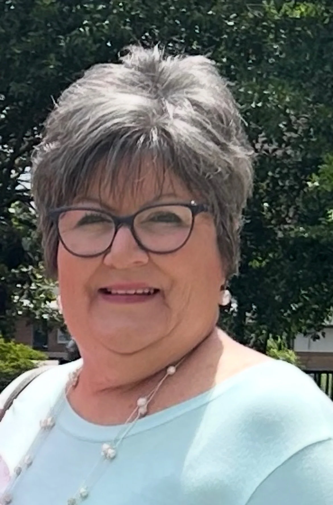 A woman with brown hair, glasses, and a pink shirt, smiling, against a gray mottled background.