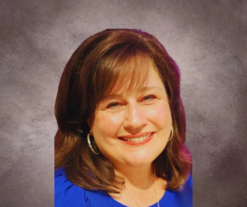 A young woman with long, wavy light brown hair, smiling, wearing a maroon shirt, standing outdoors in a wooded area.